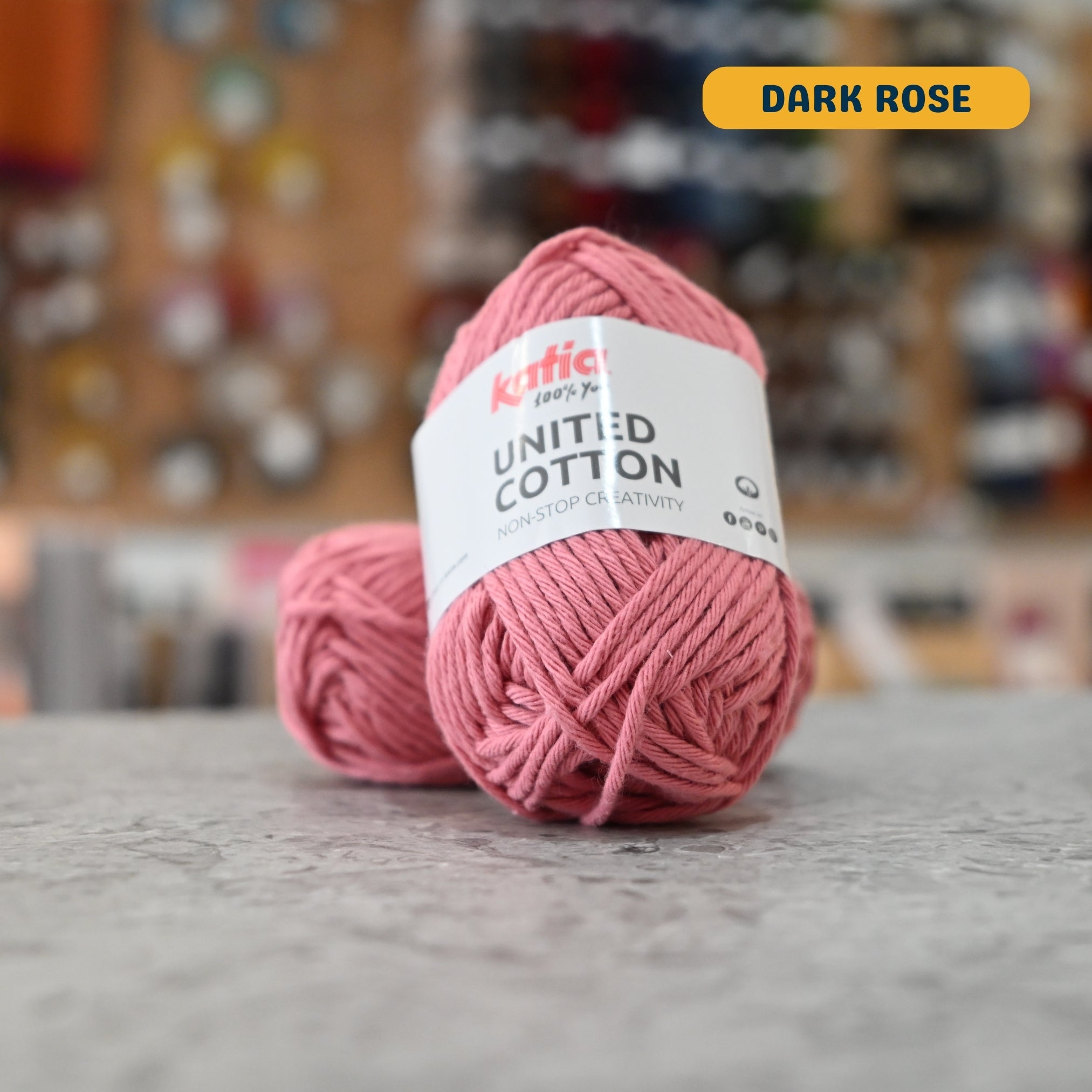 25g pink (dark rose) cotton DK yarn, 'Katia United Cotton' label visible, craft shop background (blurred)