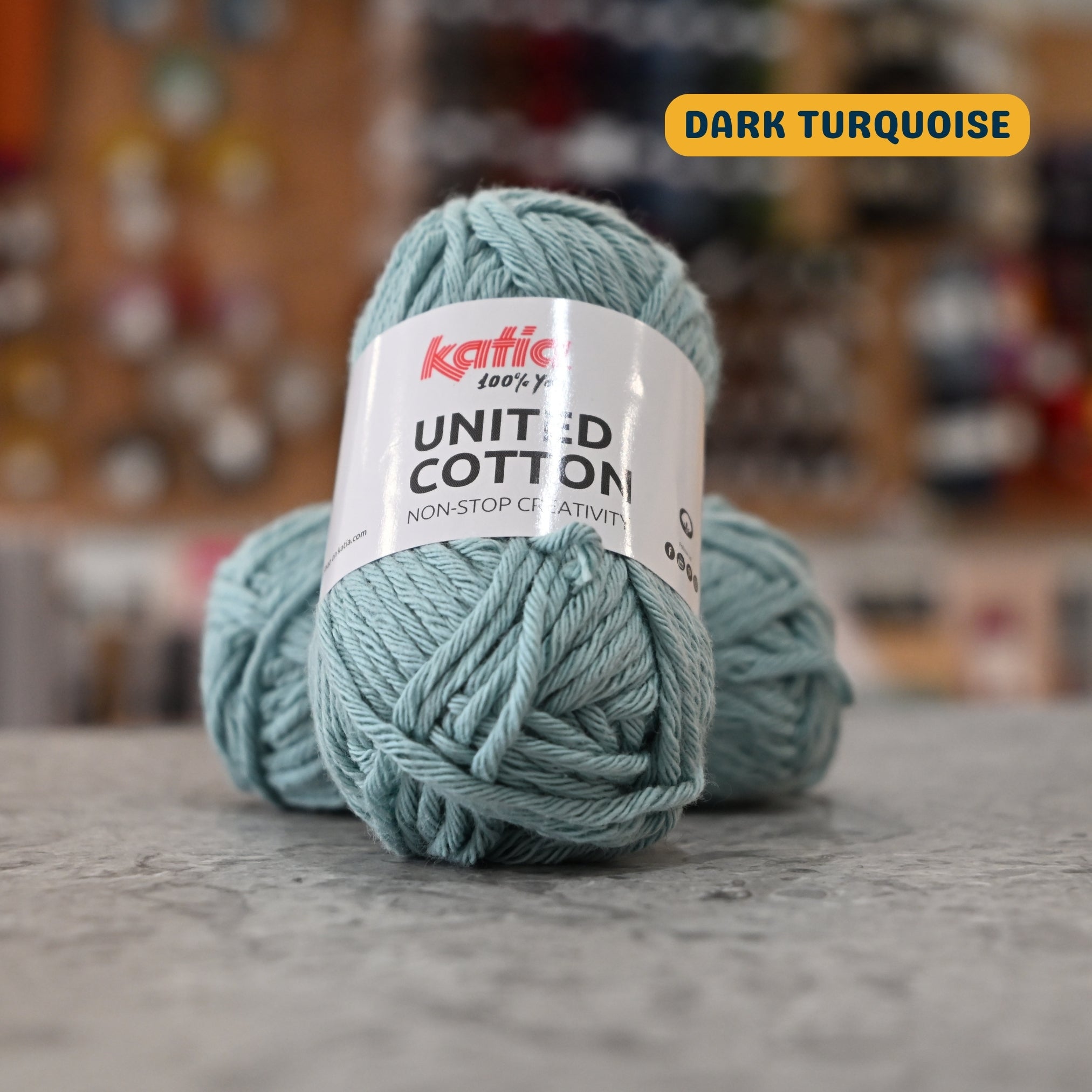 25g light blue-grey (dark turquoise) cotton DK yarn, 'Katia United Cotton' label visible, craft shop background (blurred)