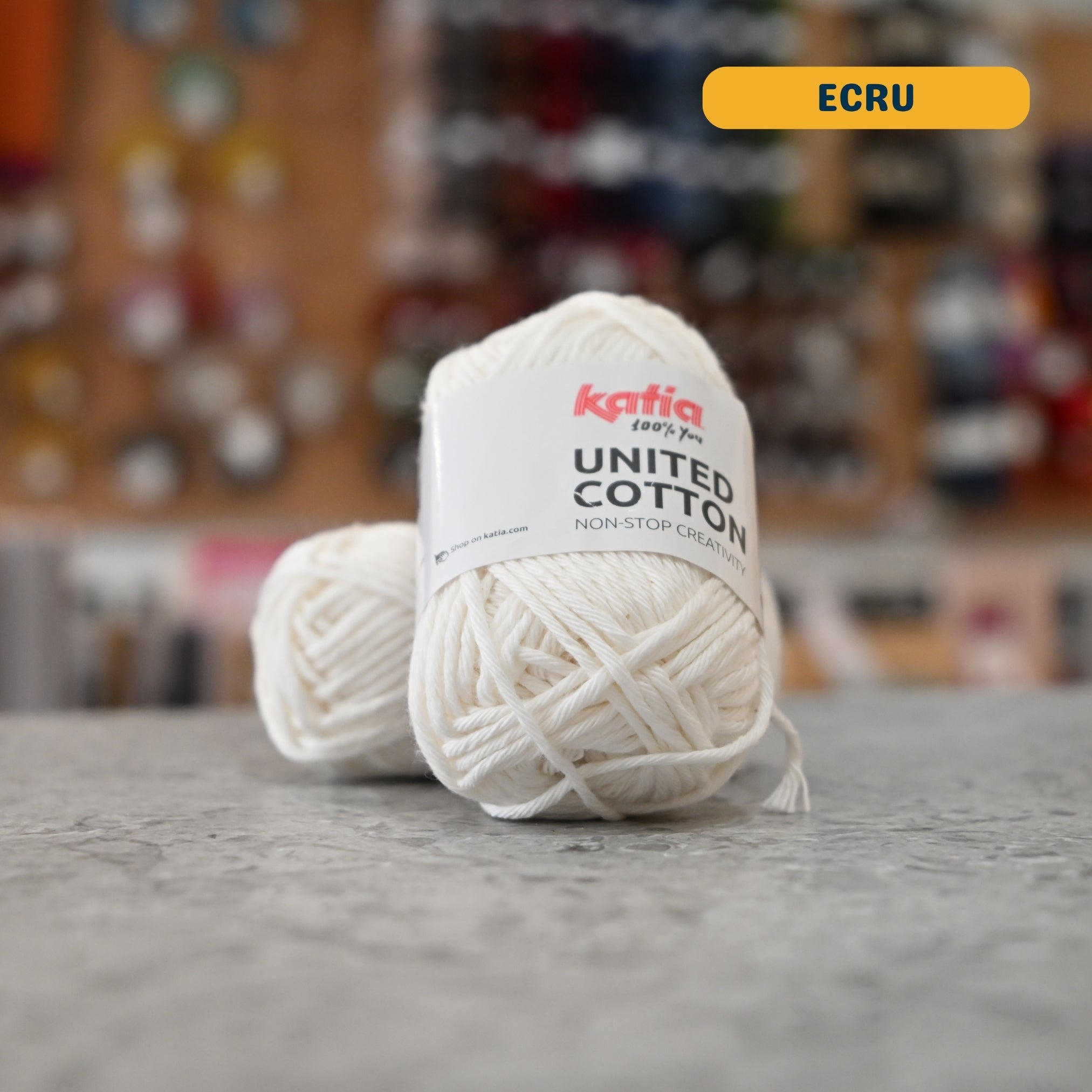 25g natural white (ecru) cotton DK yarn, 'Katia United Cotton' label visible, craft shop background (blurred)