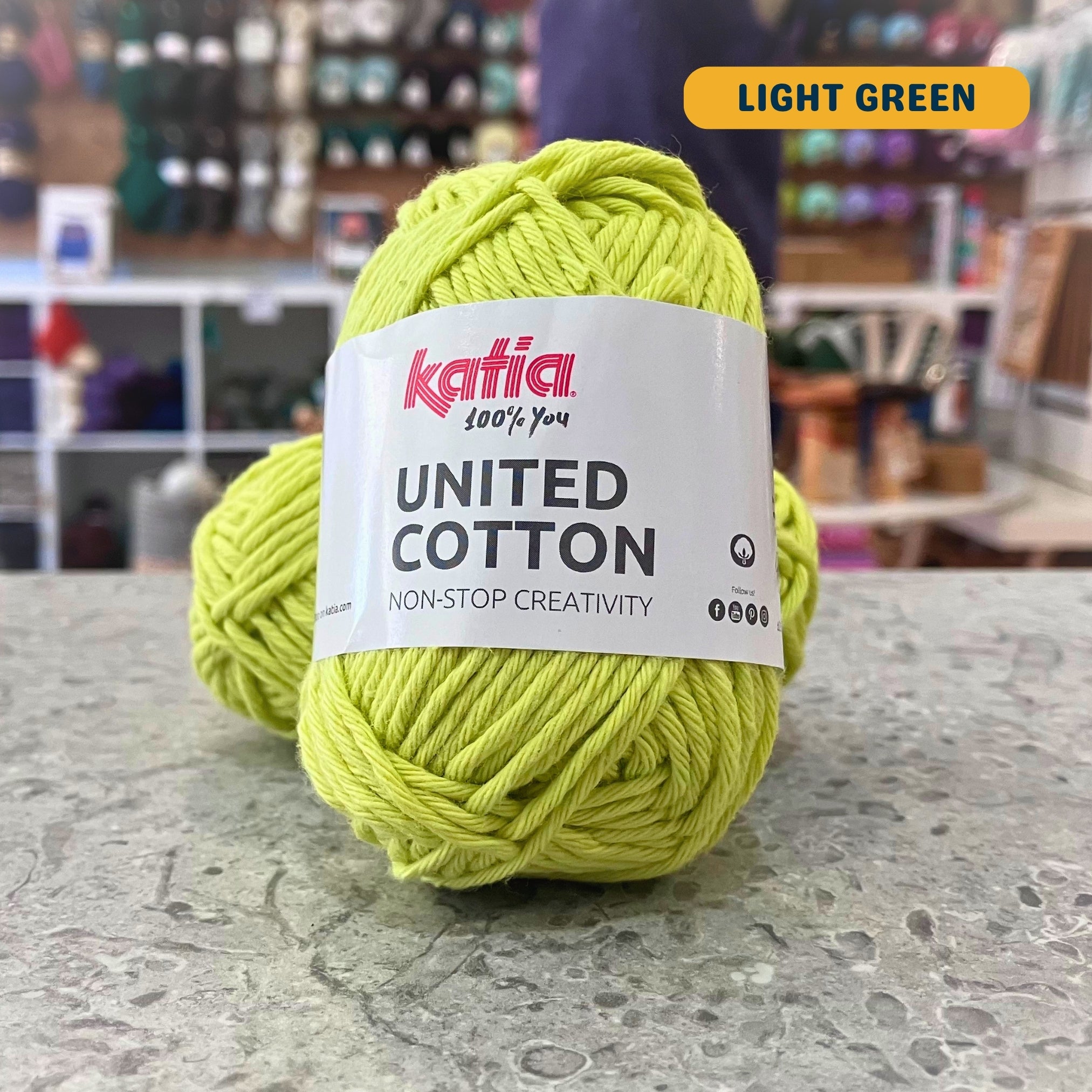 25g yellow-green (light green) cotton DK yarn, 'Katia United Cotton' label visible, craft shop background (blurred)