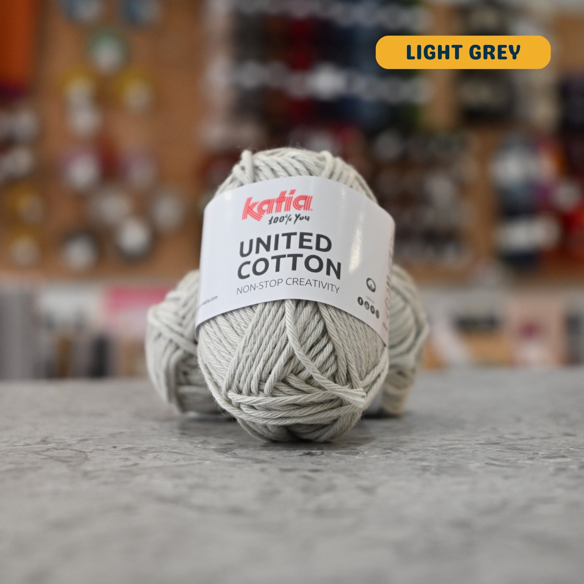 25g light grey cotton DK yarn, 'Katia United Cotton' label visible, craft shop background (blurred)