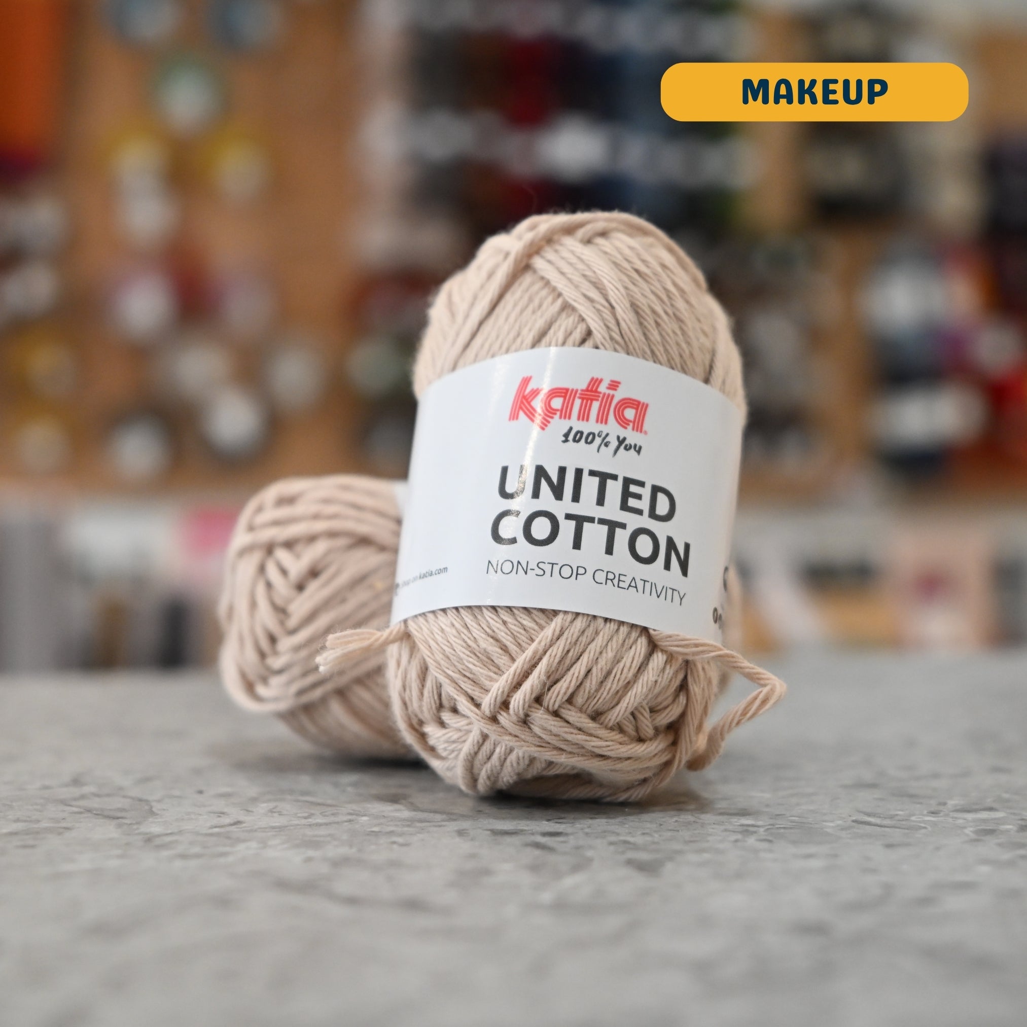 25g beige (makeup) cotton DK yarn, 'Katia United Cotton' label visible, craft shop background (blurred)
