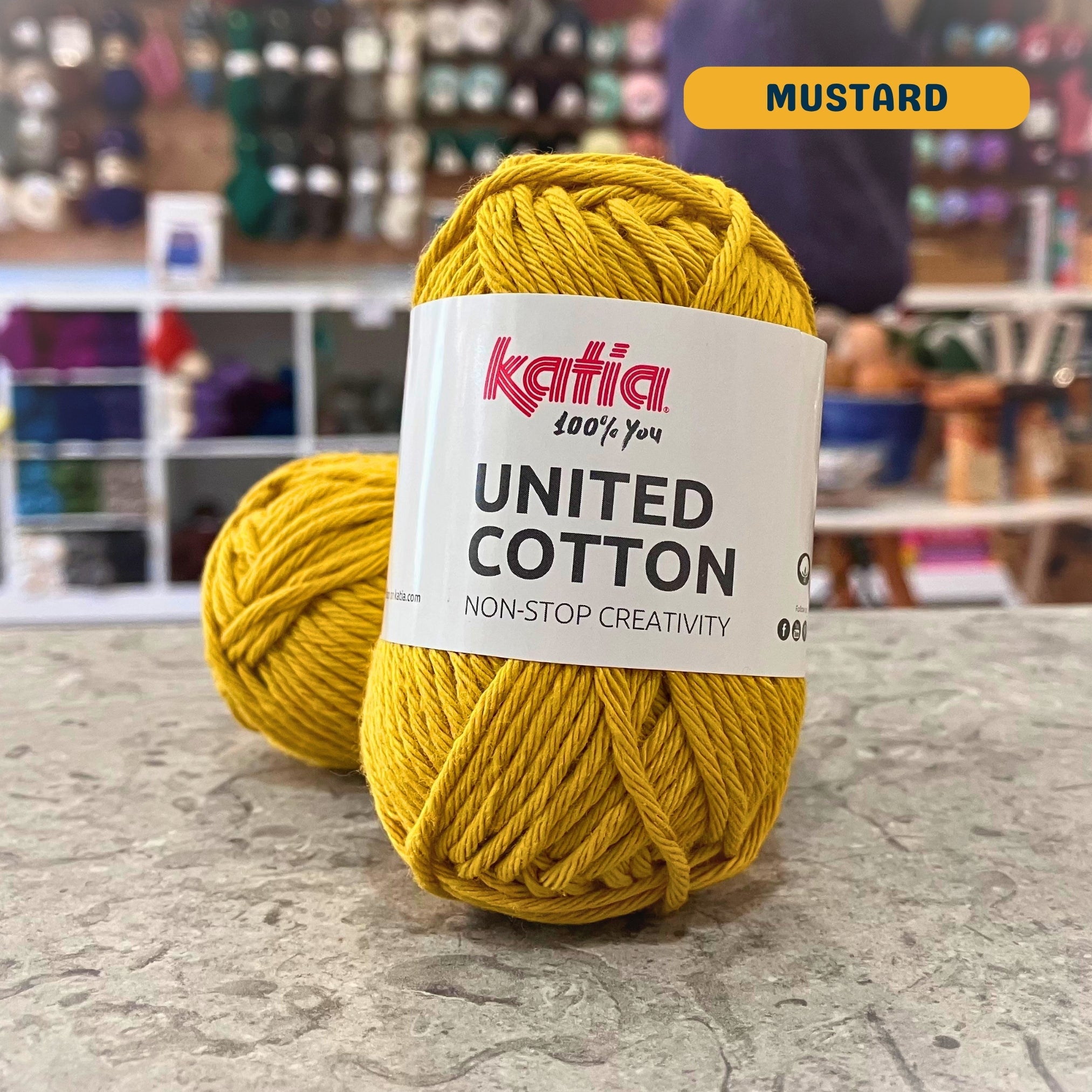 25g yellow (mustard) cotton DK yarn, 'Katia United Cotton' label visible, craft shop background (blurred)