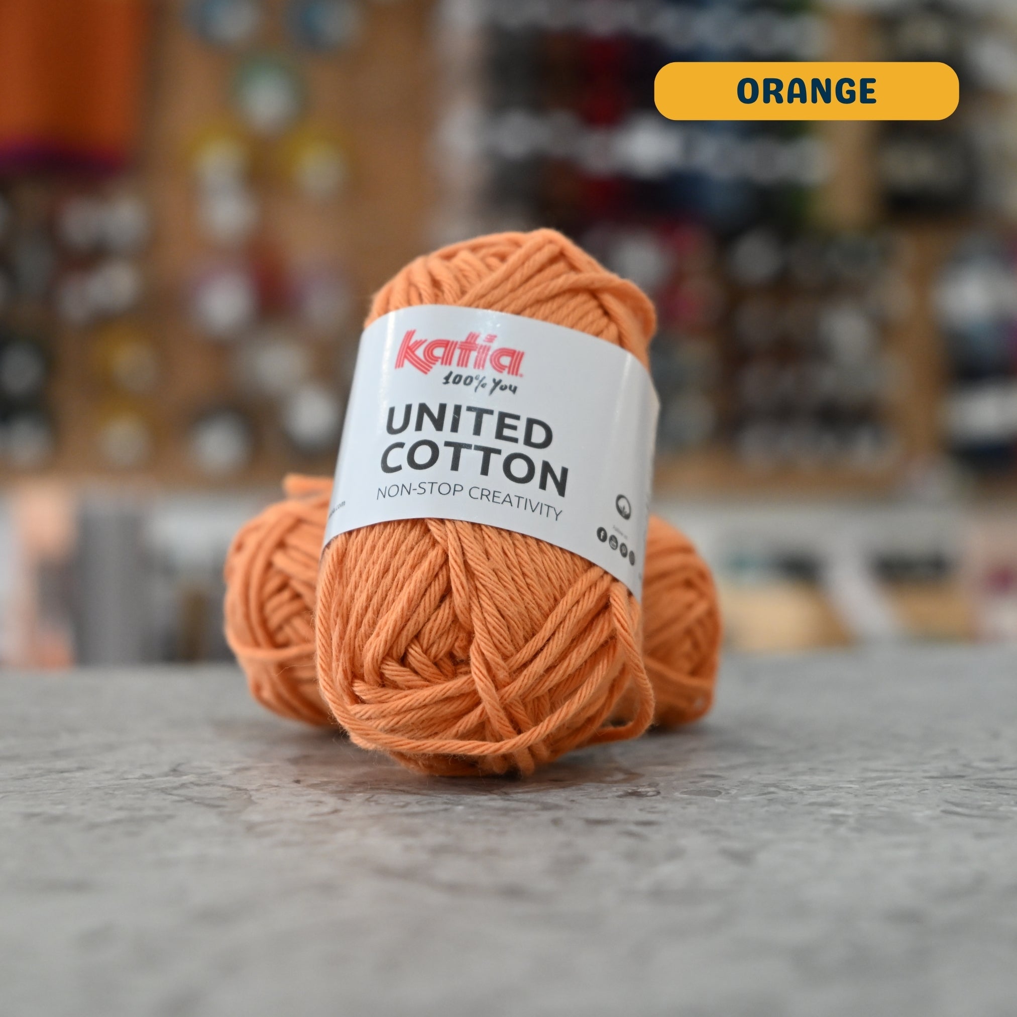 25g orange cotton DK yarn, 'Katia United Cotton' label visible, craft shop background (blurred)