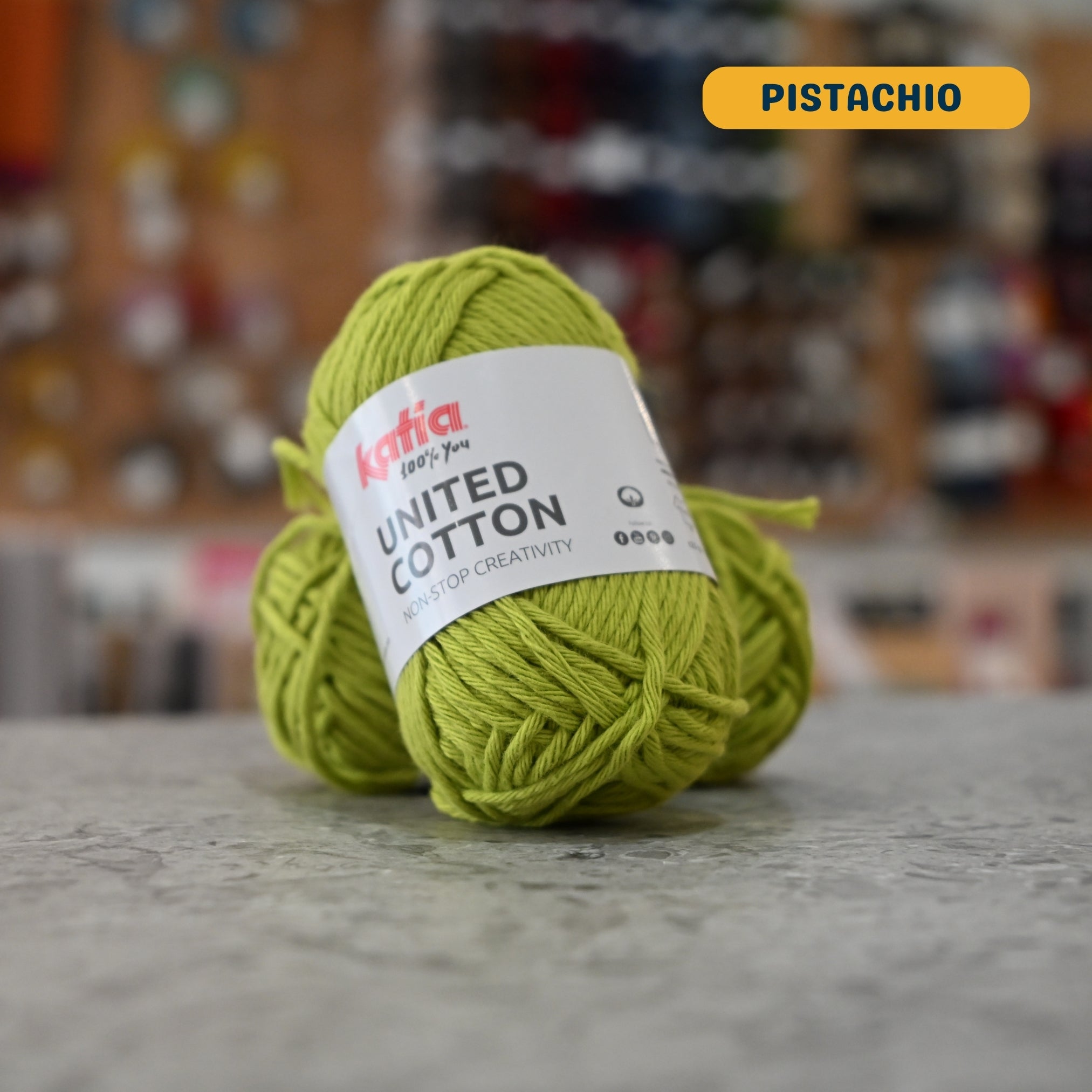 25g bright green (pistachio) cotton DK yarn, 'Katia United Cotton' label visible, craft shop background (blurred)
