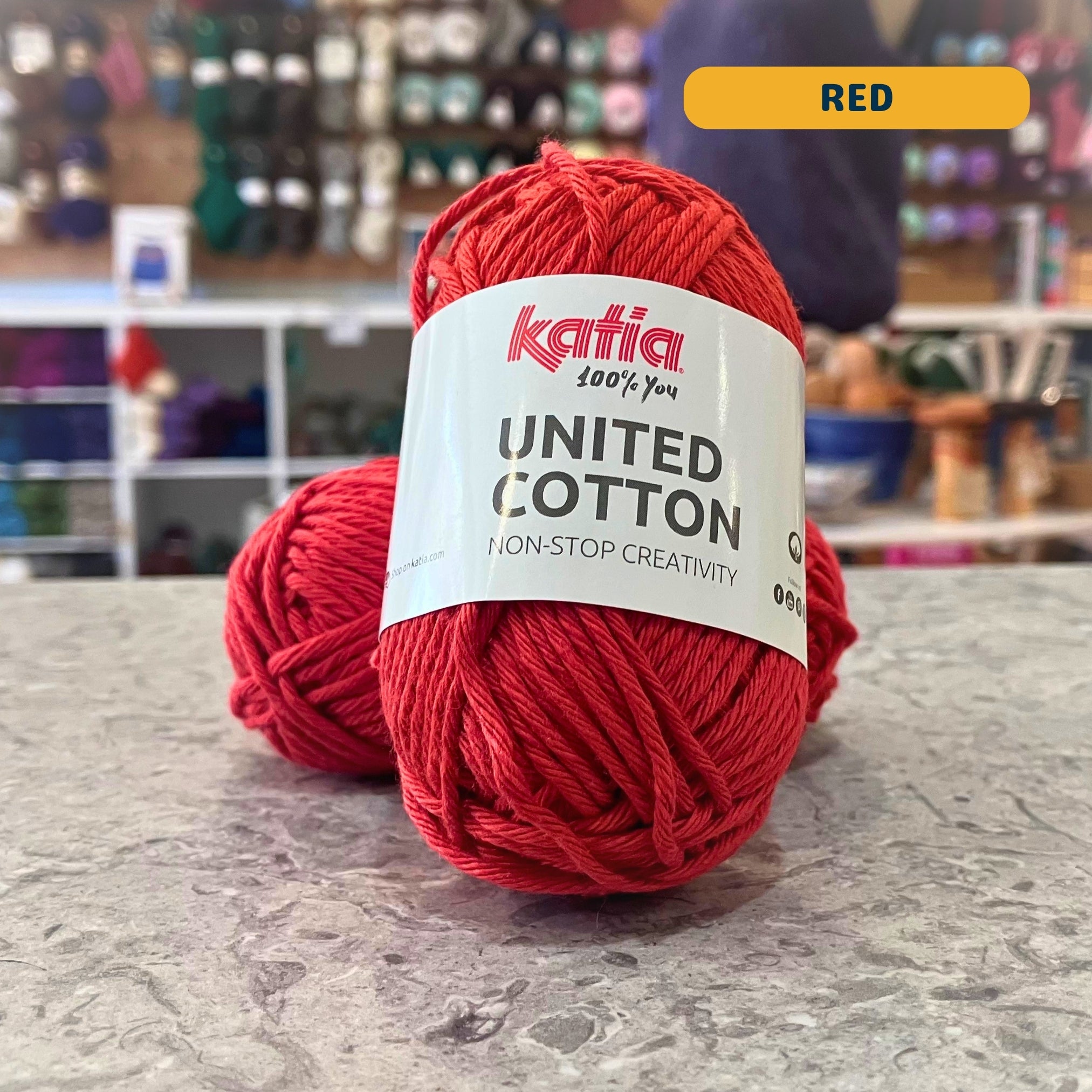 25g dark orange-red (red) cotton DK yarn, 'Katia United Cotton' label visible, craft shop background (blurred)