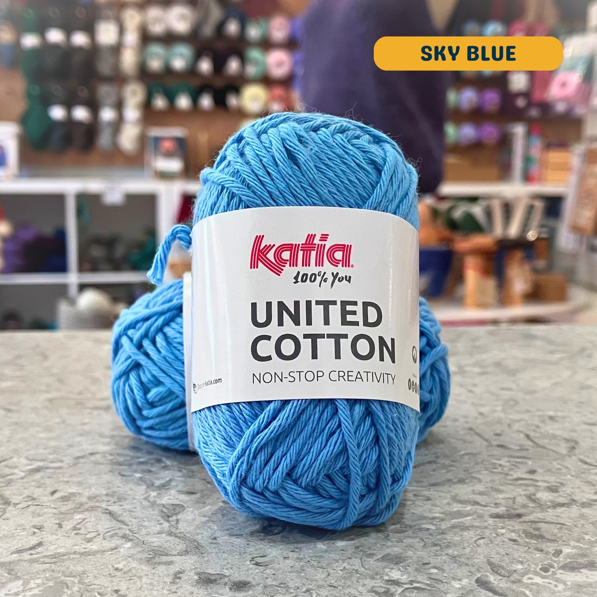 25g bright blue (sky blue) cotton DK yarn, 'Katia United Cotton' label visible, craft shop background (blurred)