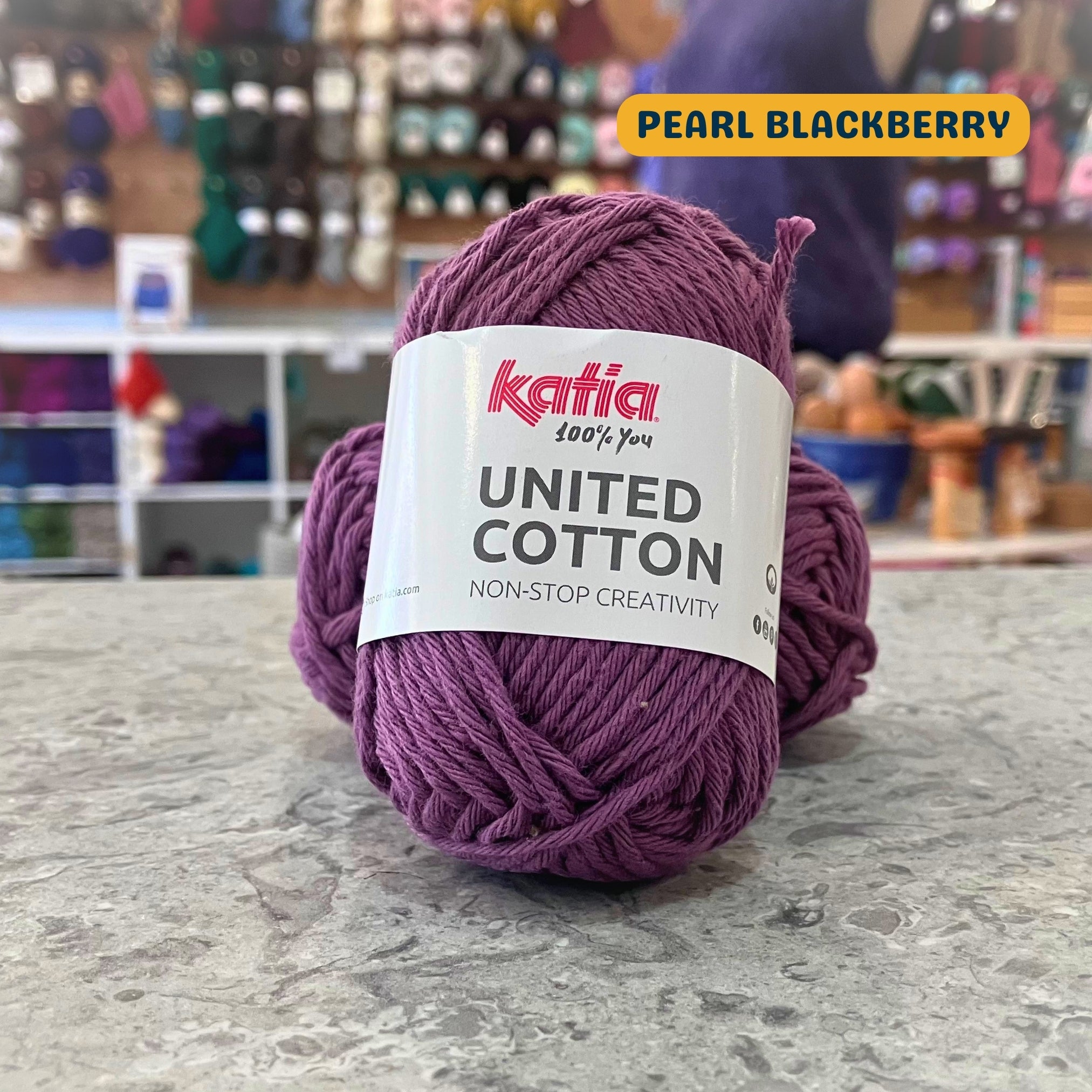 25g muted dark purple (pearl blackberry) cotton DK yarn, 'Katia United Cotton' label visible, craft shop background (blurred)