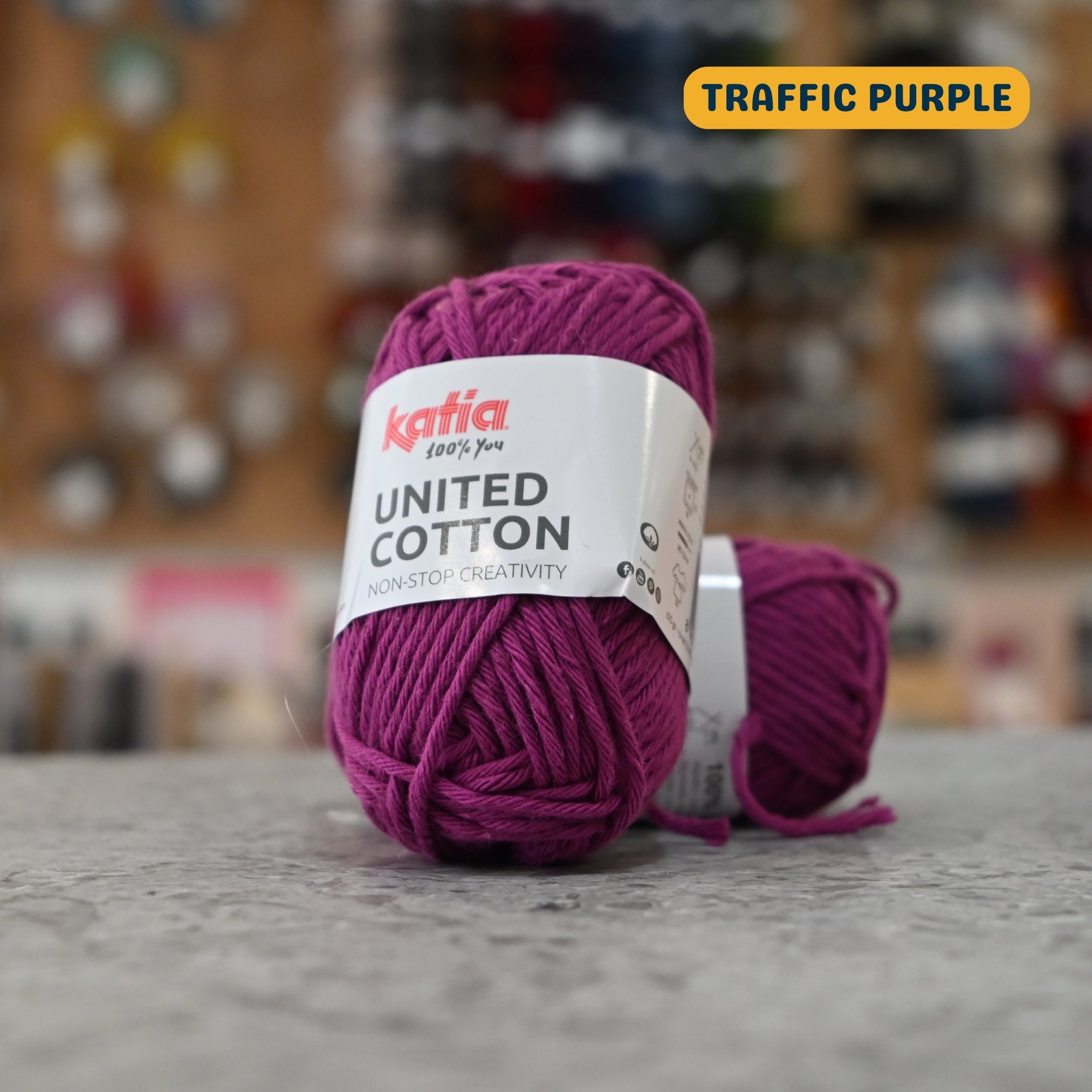 25g dark magenta (traffic purple) cotton DK yarn, 'Katia United Cotton' label visible, craft shop background (blurred)