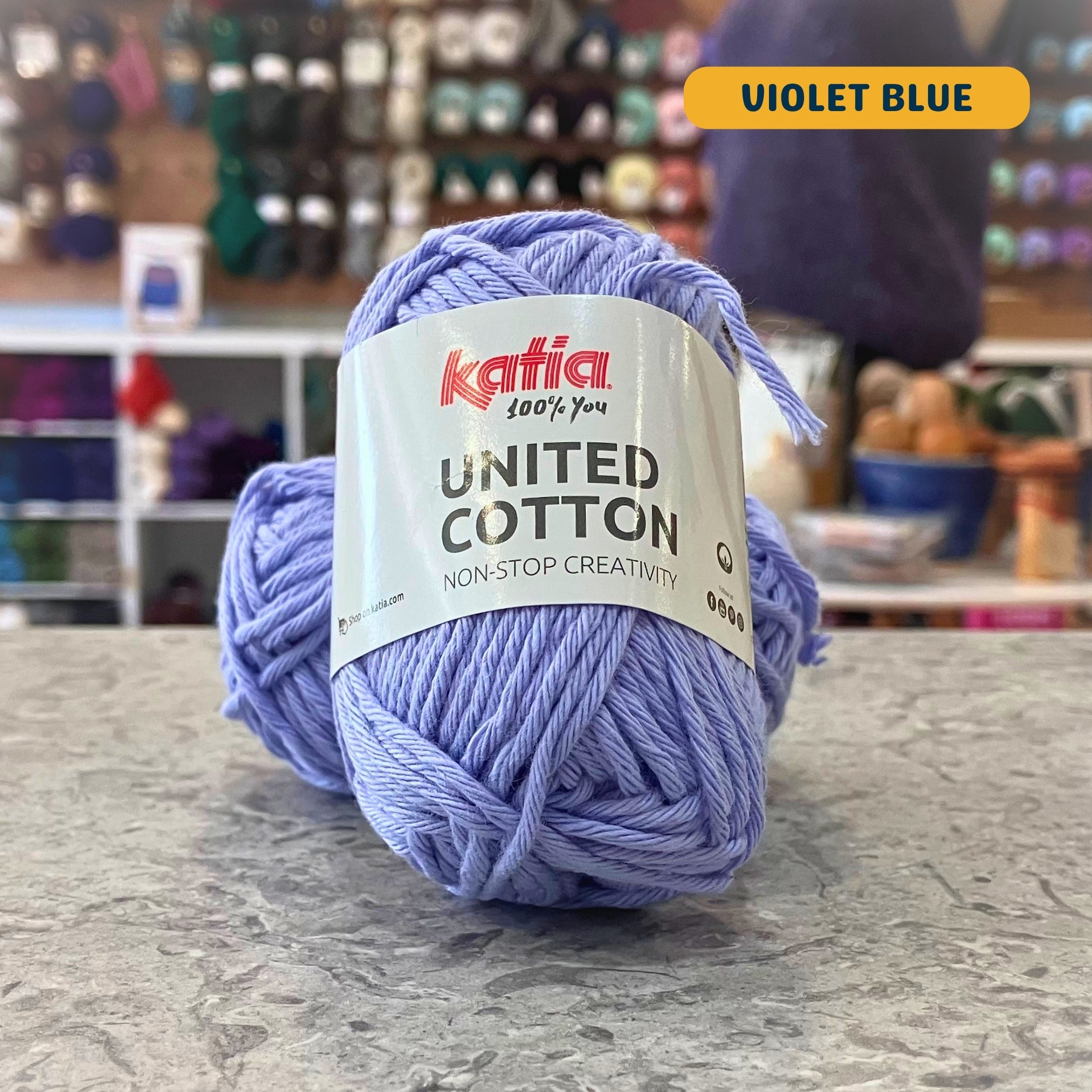 25g light purple (violet blue) cotton DK yarn, 'Katia United Cotton' label visible, craft shop background (blurred)