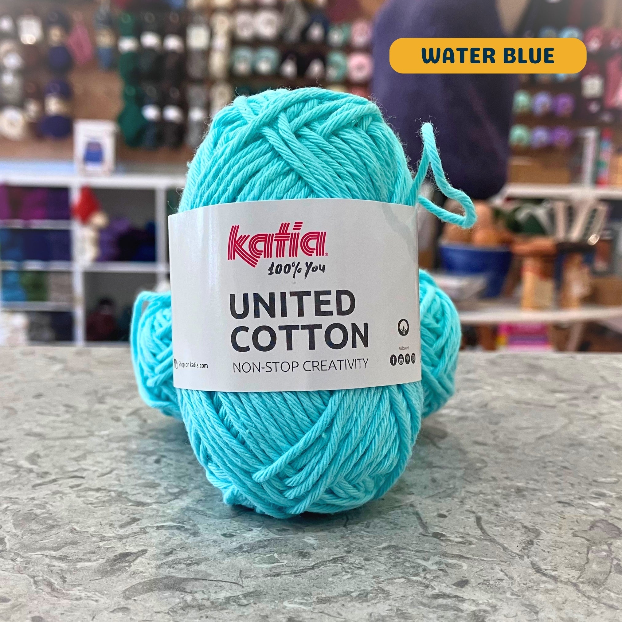 25g vibrant light teal (water blue) cotton DK yarn, 'Katia United Cotton' label visible, craft shop background (blurred)