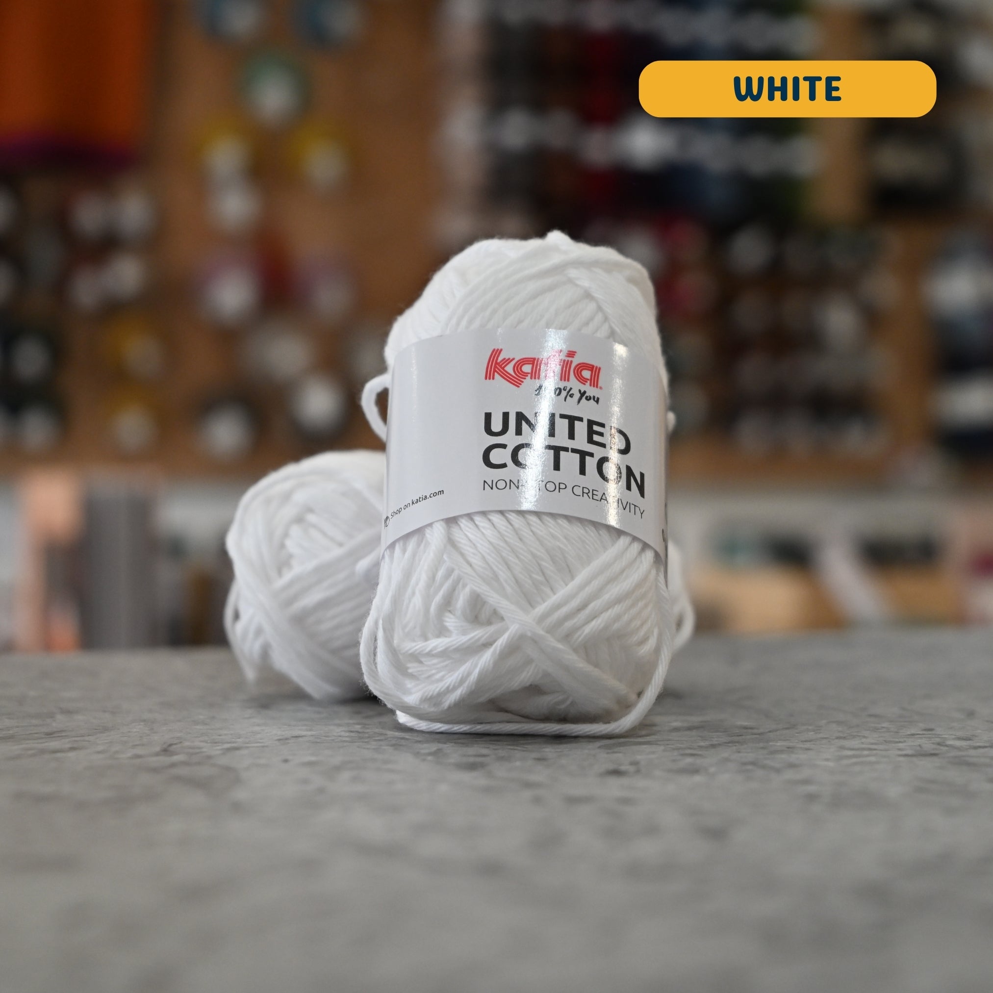 25g bleached white cotton DK yarn, 'Katia United Cotton' label visible, craft shop background (blurred)