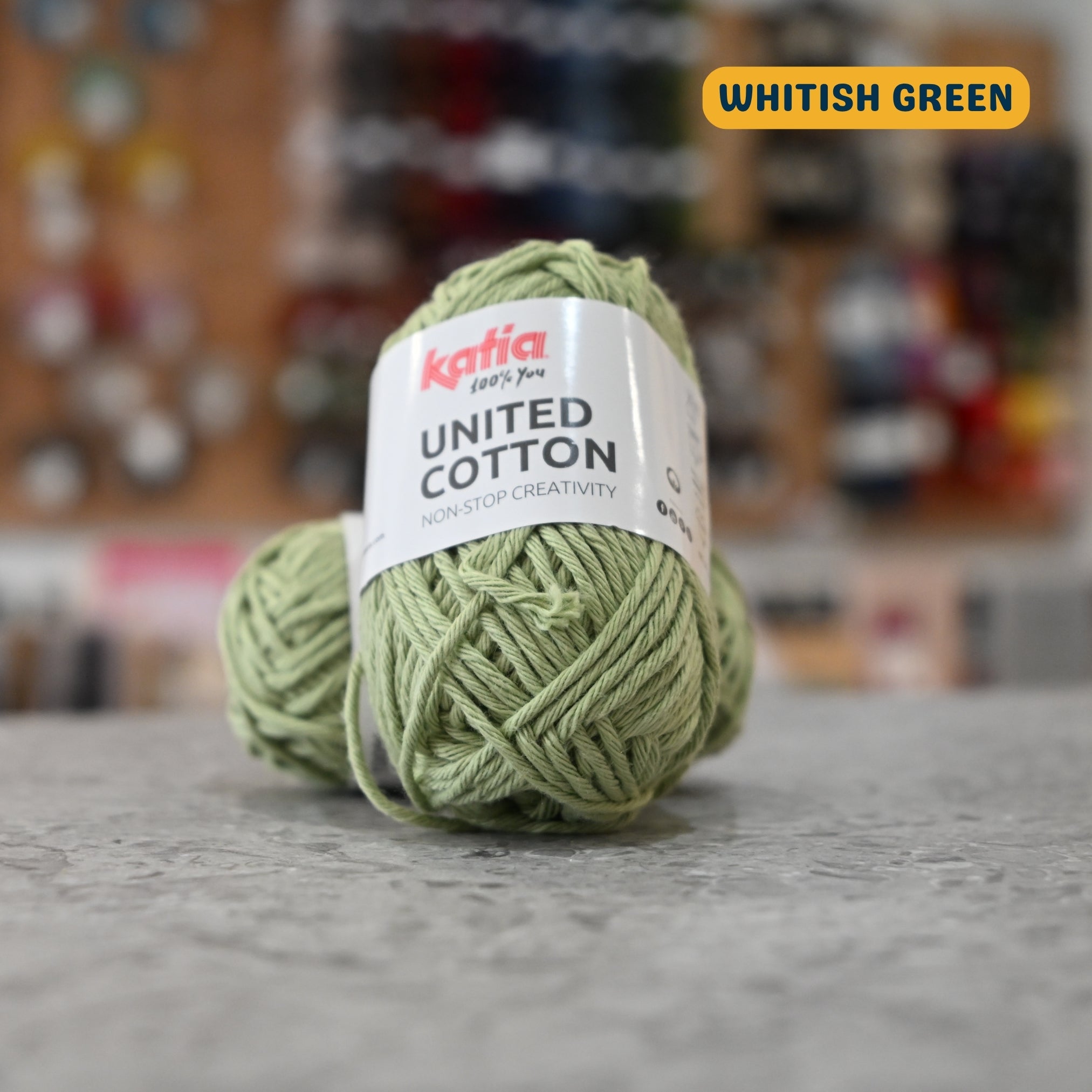 25g sage (whitish green) cotton DK yarn, 'Katia United Cotton' label visible, craft shop background (blurred)