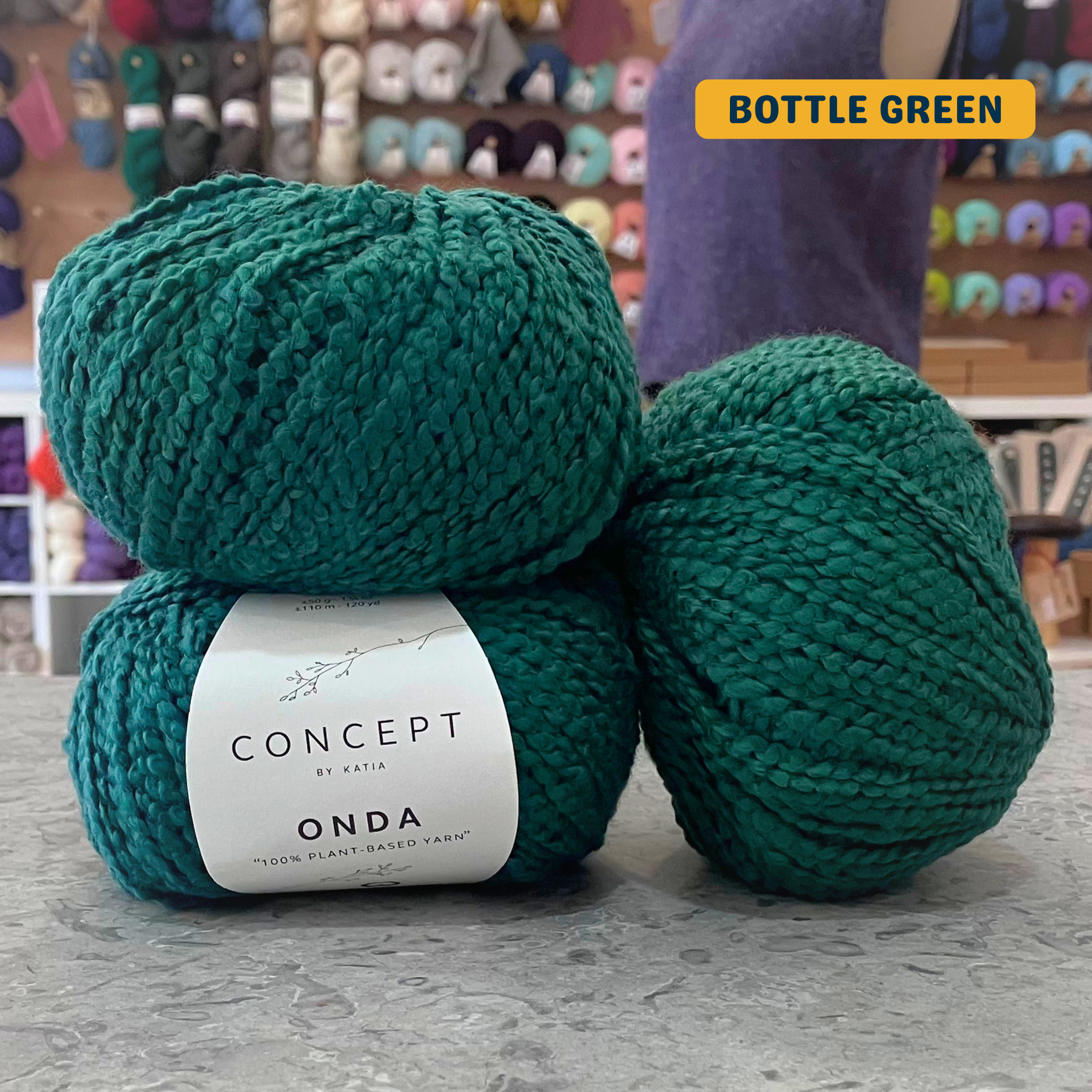 50g blue-green (bottle green) boucle textured cotton worsted yarn, 'Concept by Katia Odna 100% Plant-based Yarn' label visible, craft shop background (blurred)