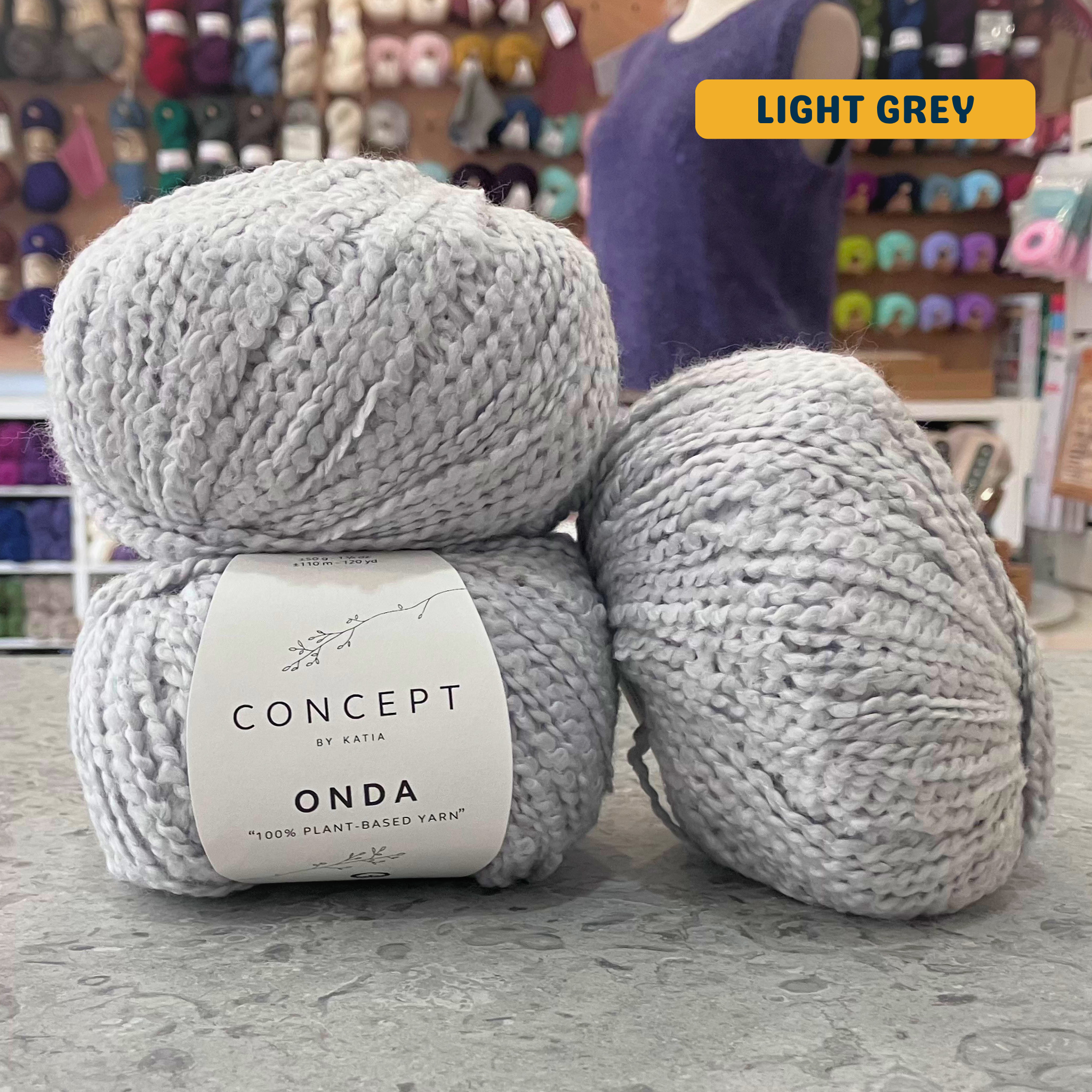 50g light grey boucle textured cotton worsted yarn, 'Concept by Katia Odna 100% Plant-based Yarn' label visible, craft shop background (blurred)