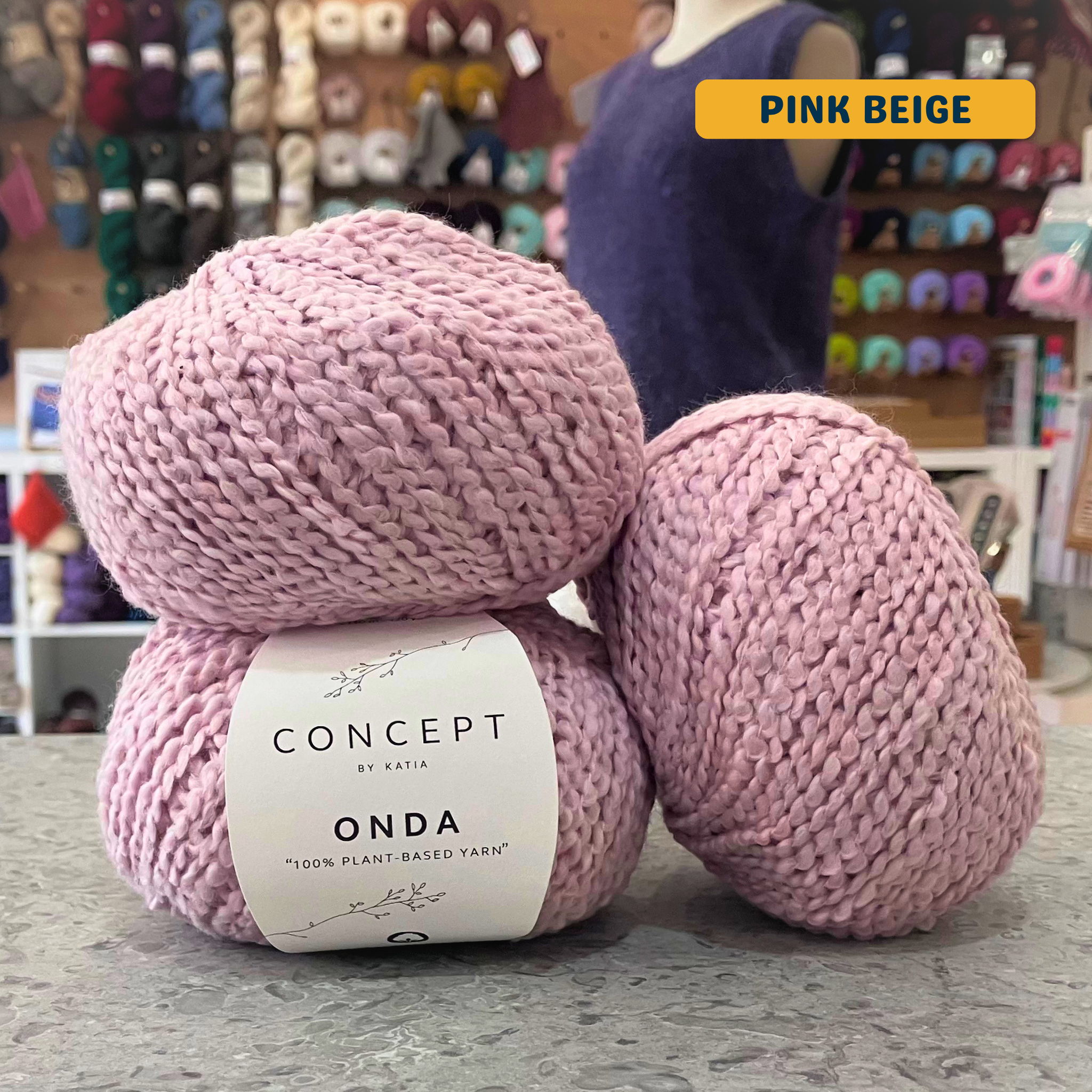 50g muted pink (pink beige) boucle textured cotton worsted yarn, 'Concept by Katia Odna 100% Plant-based Yarn' label visible, craft shop background (blurred)