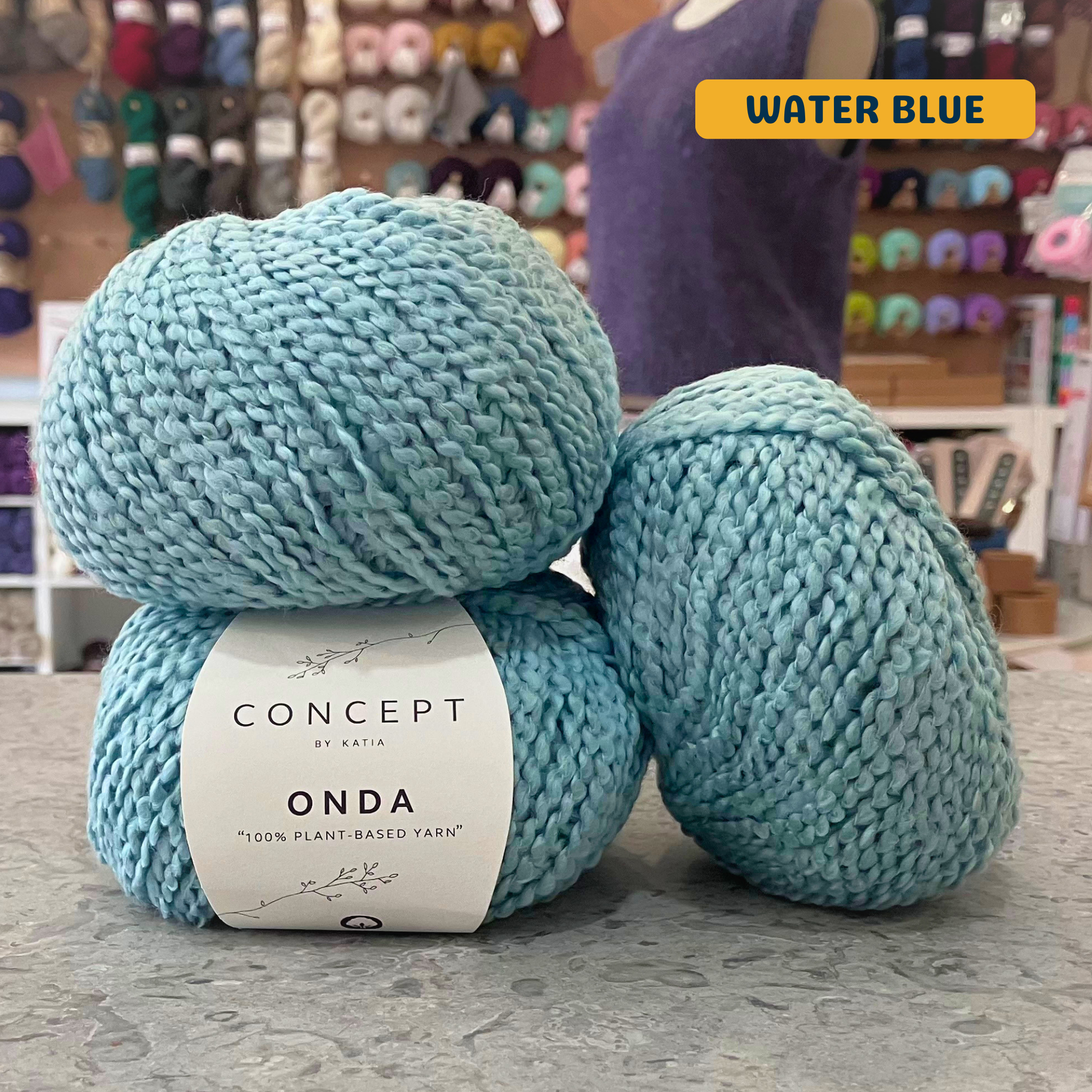 50g light blue (water blue) boucle textured cotton worsted yarn, 'Concept by Katia Odna 100% Plant-based Yarn' label visible, craft shop background (blurred)