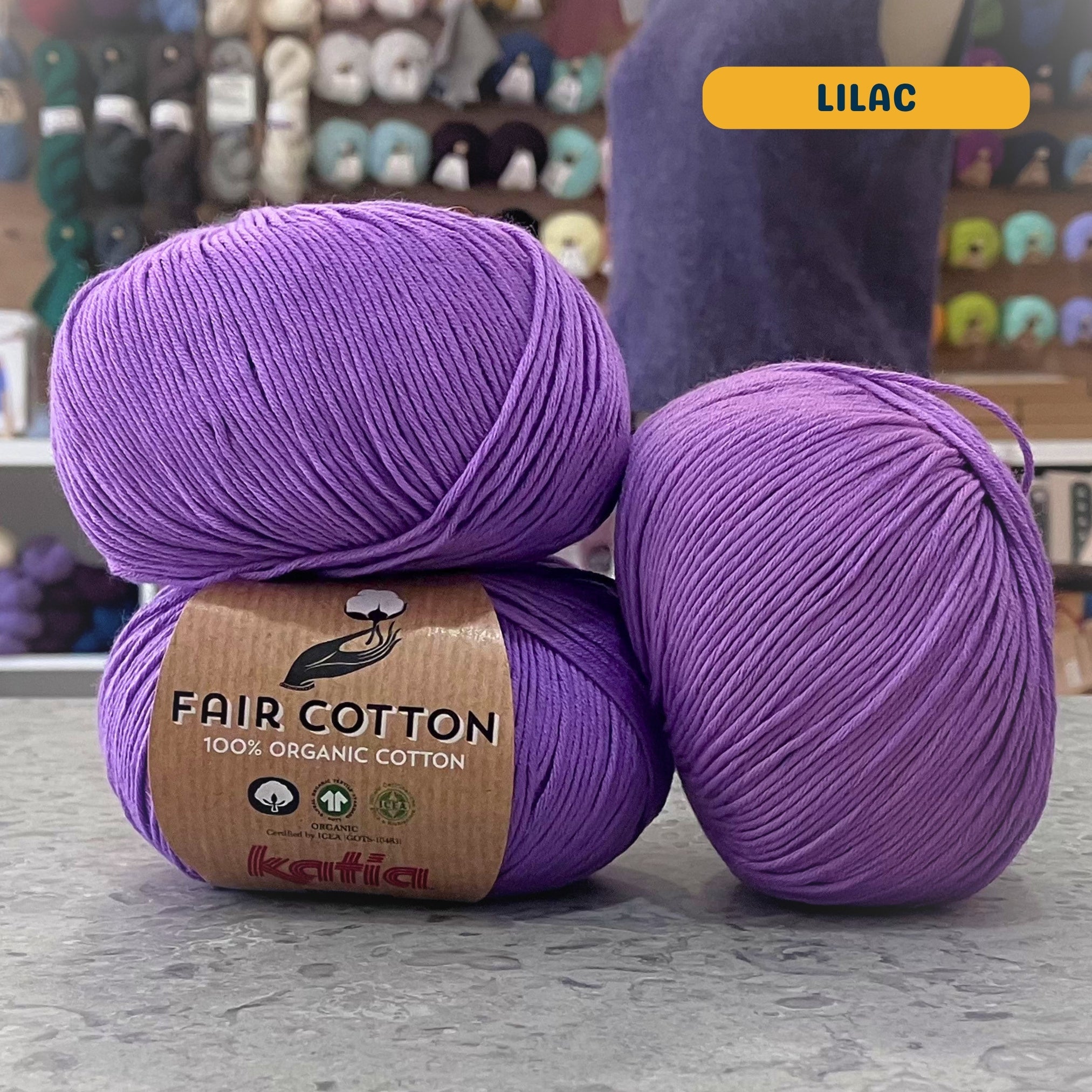 50g medium purple (lilac) organic cotton sport yarn, 'Fair Cotton 100% Organic Cotton Katia' label visible, craft shop background (blurred)