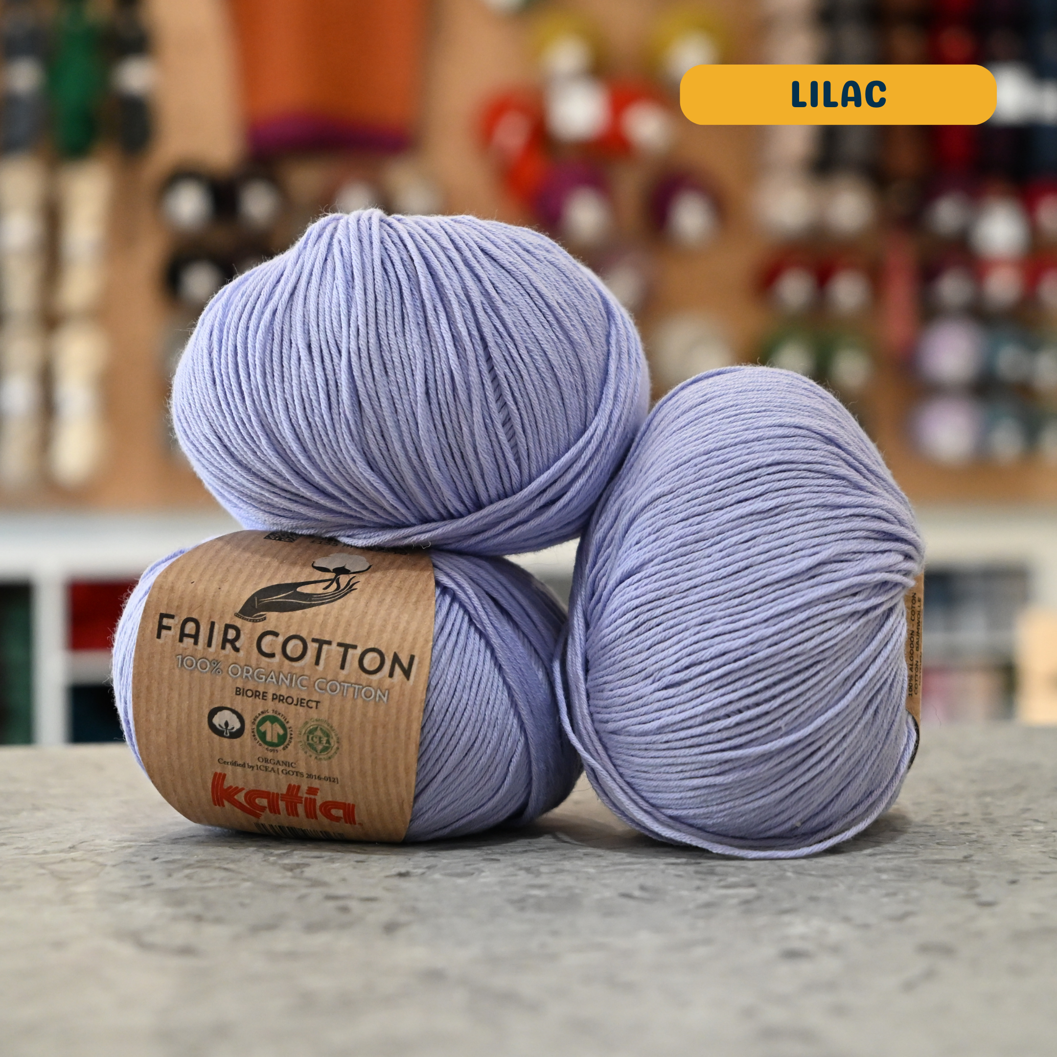 50g pale purple (lilac) organic cotton sport yarn, 'Fair Cotton 100% Organic Cotton Katia' label visible, craft shop background (blurred)