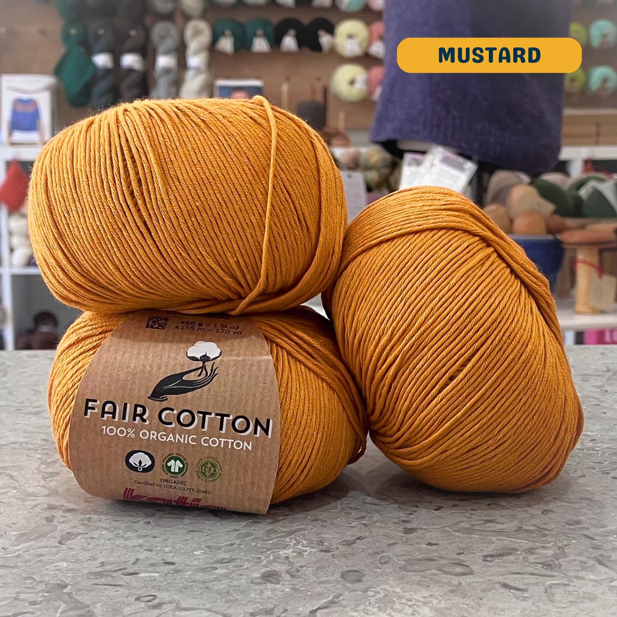 50g orange-yellow (mustard) organic cotton sport yarn, 'Fair Cotton 100% Organic Cotton Katia' label visible, craft shop background (blurred)