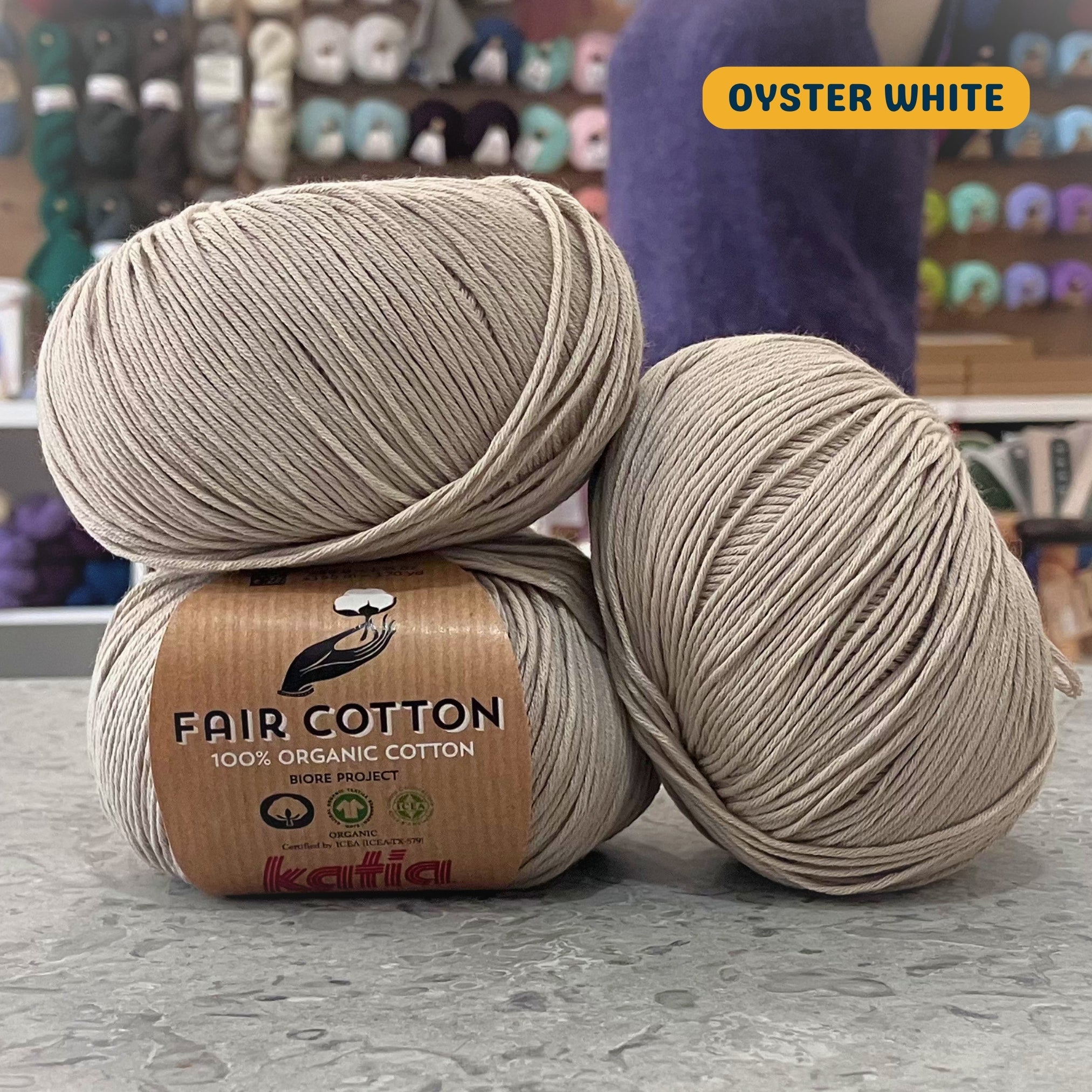 50g grey-beige (oyster white) organic cotton sport yarn, 'Fair Cotton 100% Organic Cotton Katia' label visible, craft shop background (blurred)