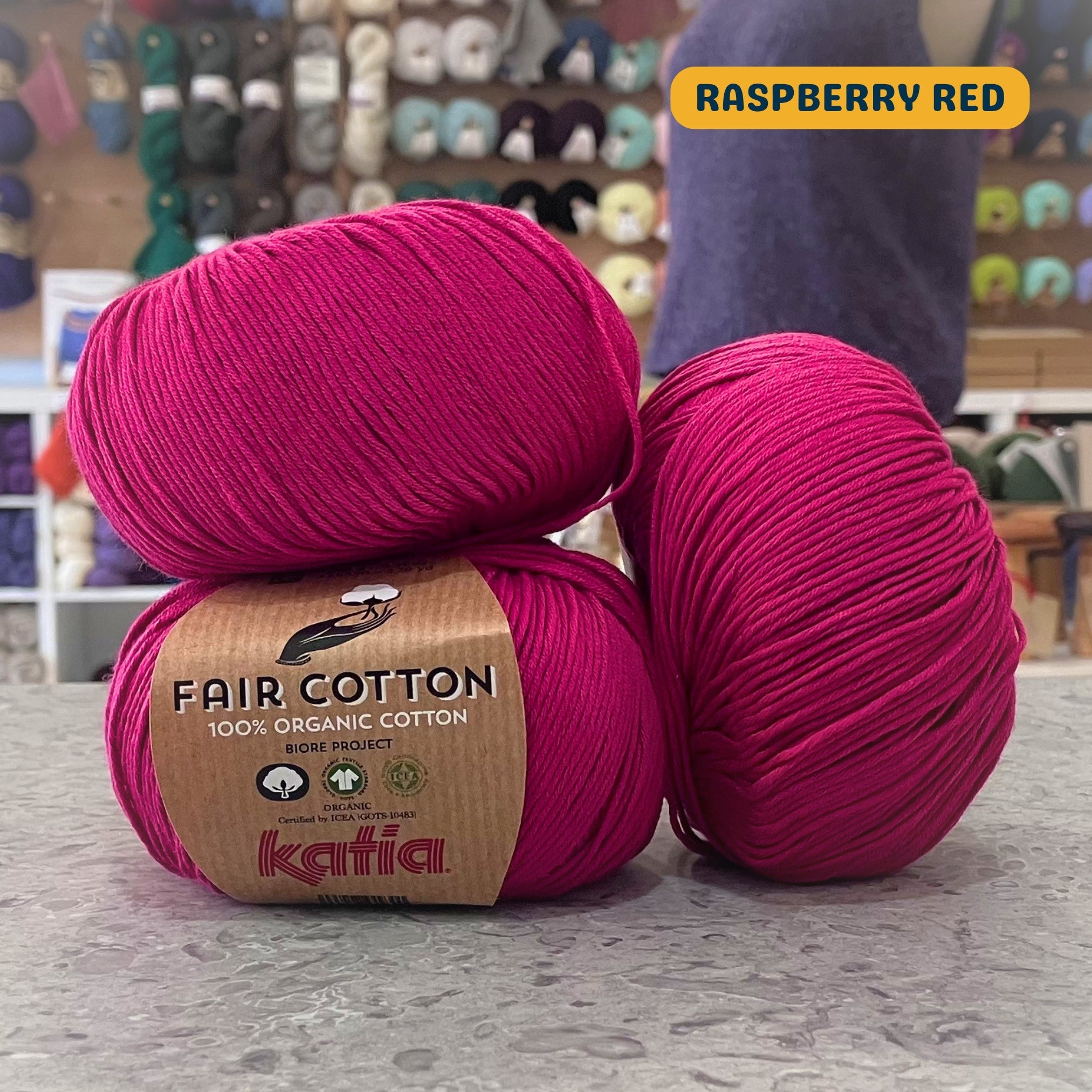 50g dark pink (raspberry red) organic cotton sport yarn, 'Fair Cotton 100% Organic Cotton Katia' label visible, craft shop background (blurred)