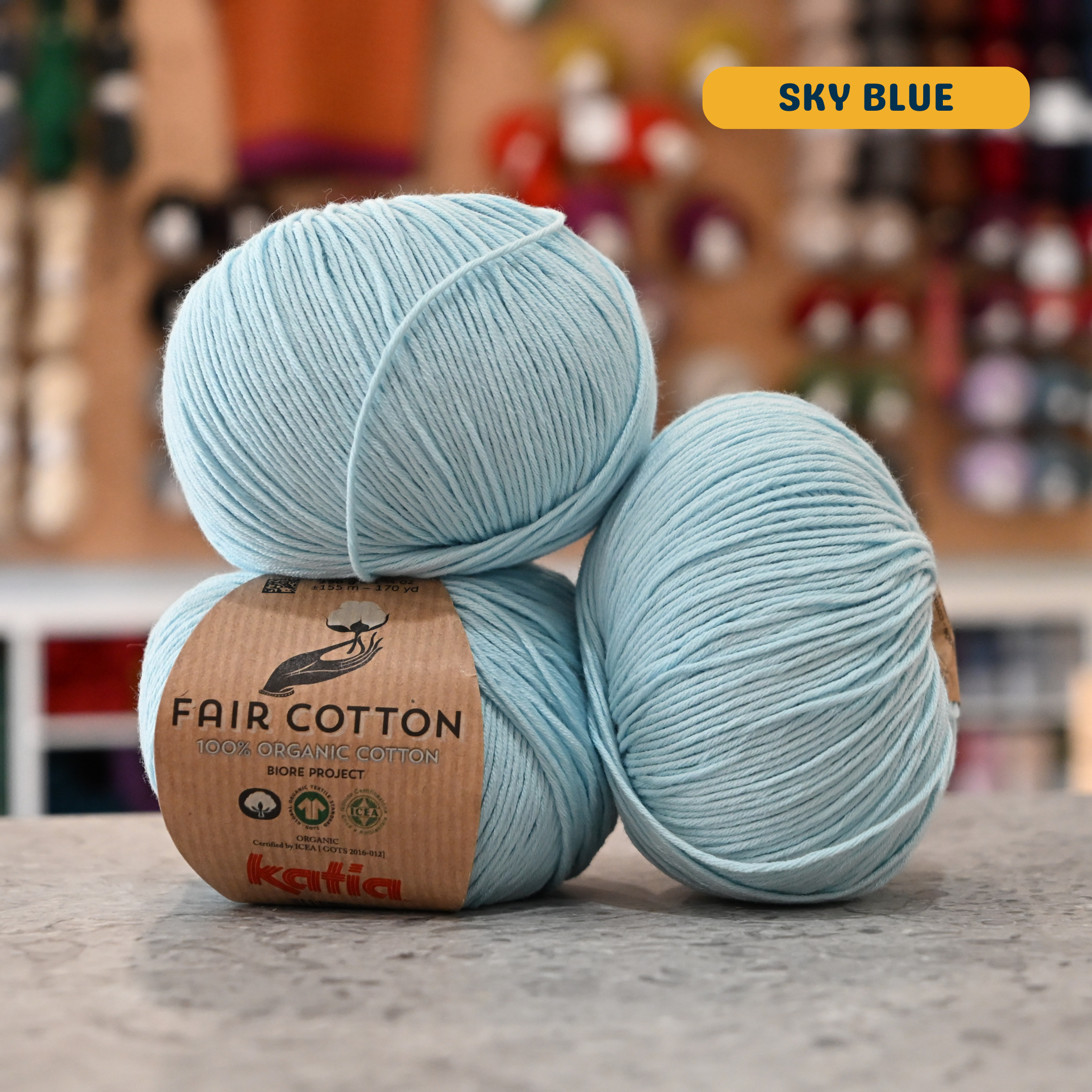 50g sky blue organic cotton sport yarn, 'Fair Cotton 100% Organic Cotton Katia' label visible, craft shop background (blurred)