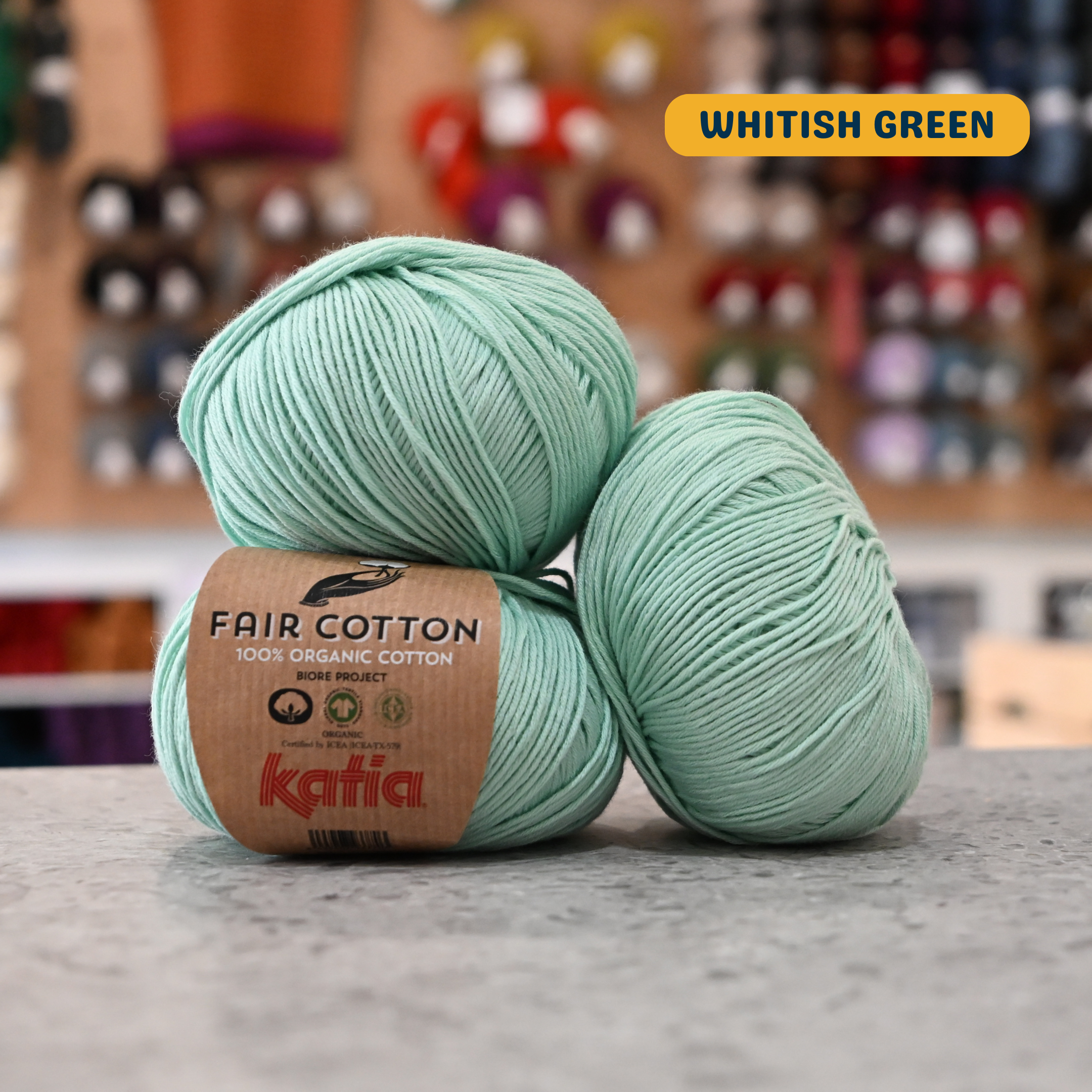 50g light teal (whitish green) organic cotton sport yarn, 'Fair Cotton 100% Organic Cotton Katia' label visible, craft shop background (blurred)