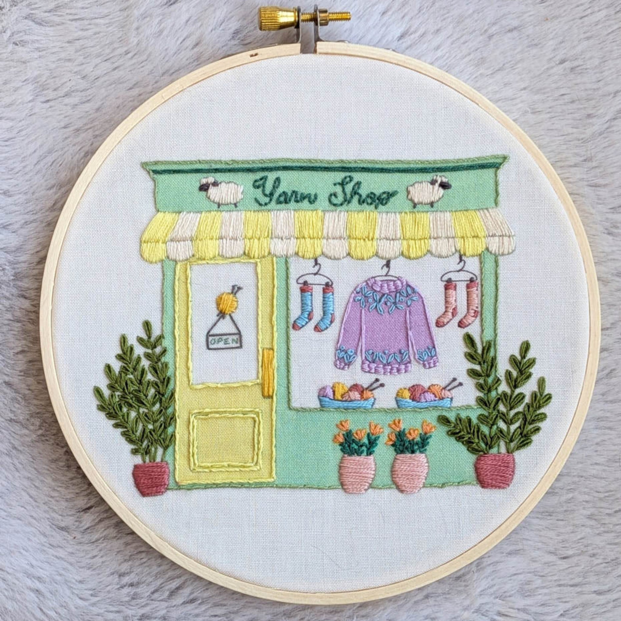 Let's Go To The Yarn Shop embroidery kit by Knitted Bliss — whimsical yarn shop scene with socks and sweaters in the window. Canadian-made.
