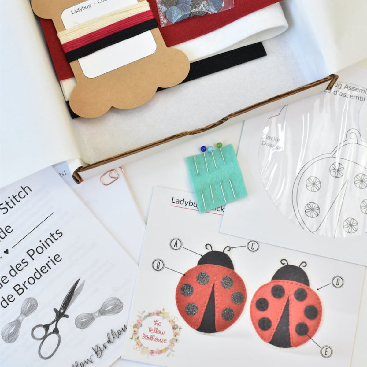 A boxed sewing or craft kit with fabric pieces, thread, pins, clear plastic templates, printed instruction sheets, and ladybug applique pieces arranged inside and around an open cardboard box.