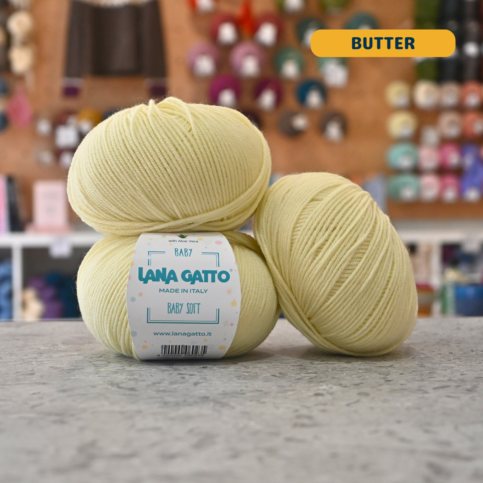 50g butter yellow extrafine merino aloe vera sport yarn, 'Lana Gatto Baby Soft' label visible, craft shop background (blurred)