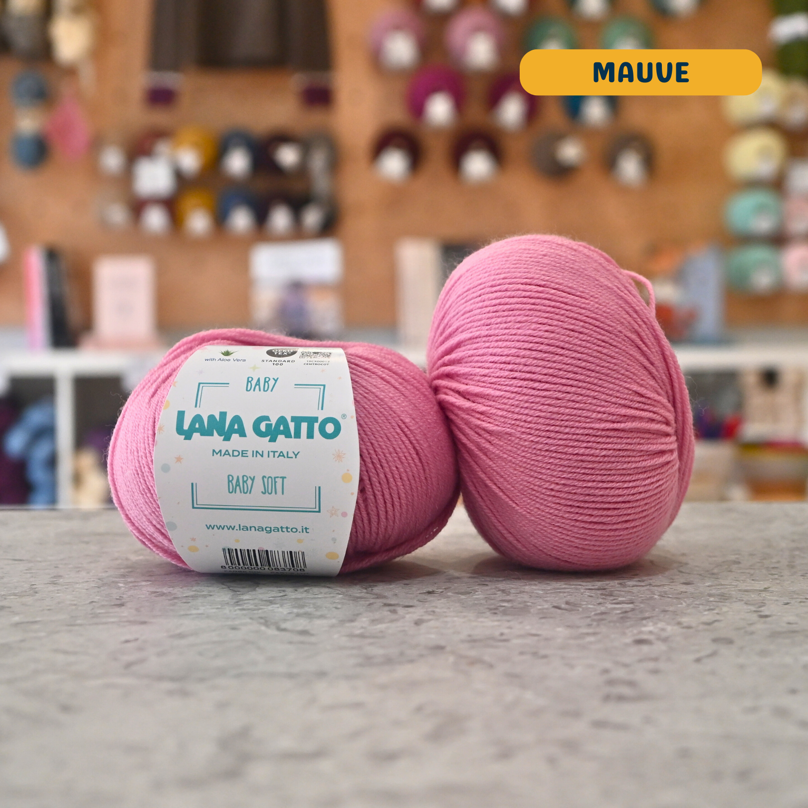 50g pink (mauve) extrafine merino aloe vera sport yarn, 'Lana Gatto Baby Soft' label visible, craft shop background (blurred)