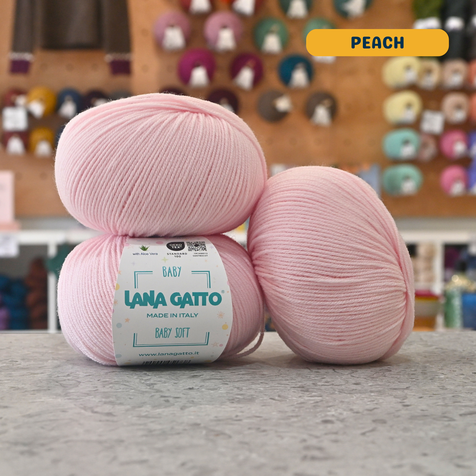 50g pale pink (peach) extrafine merino aloe vera sport yarn, 'Lana Gatto Baby Soft' label visible, craft shop background (blurred)