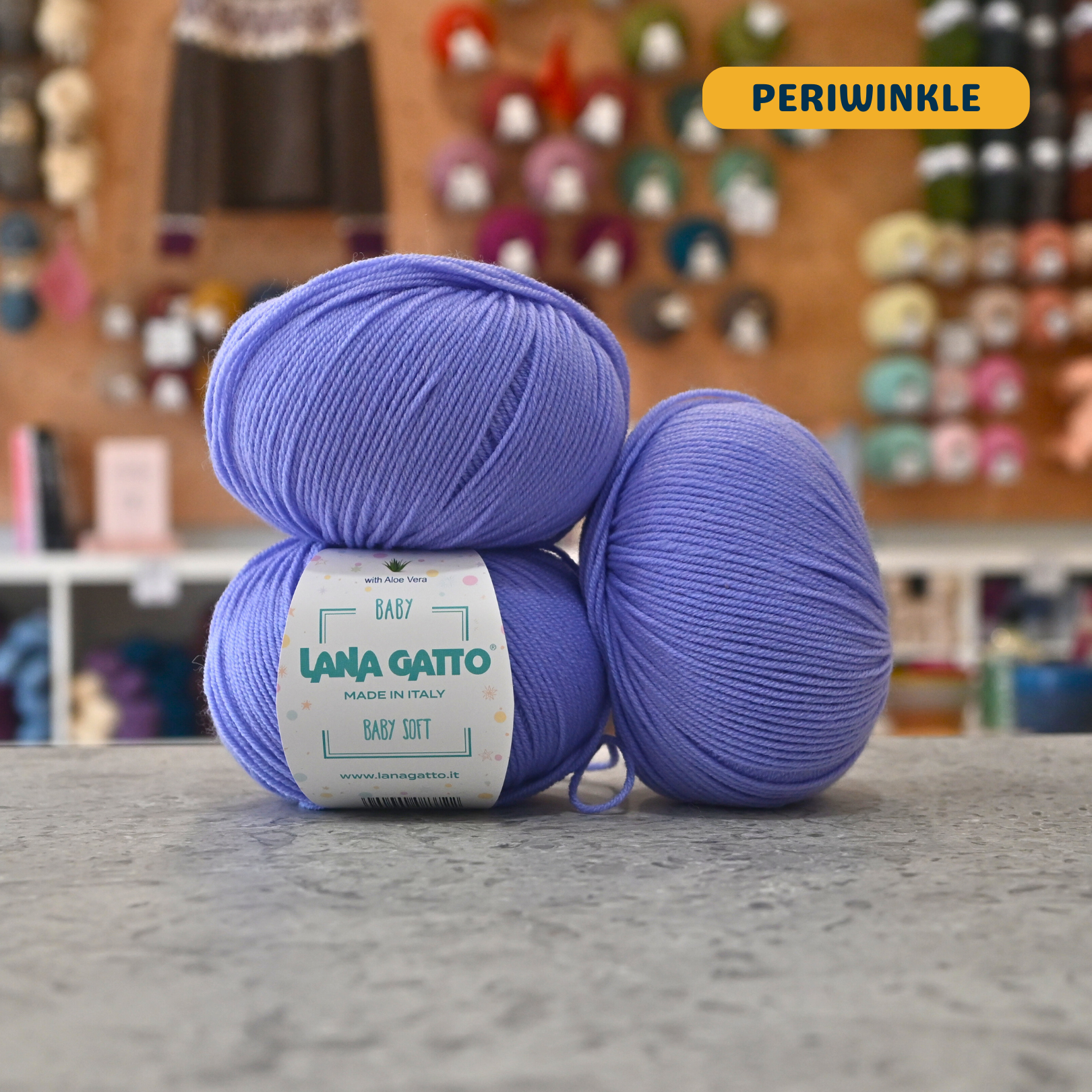 50g blue-purple (periwinkle) extrafine merino aloe vera sport yarn, 'Lana Gatto Baby Soft' label visible, craft shop background (blurred)