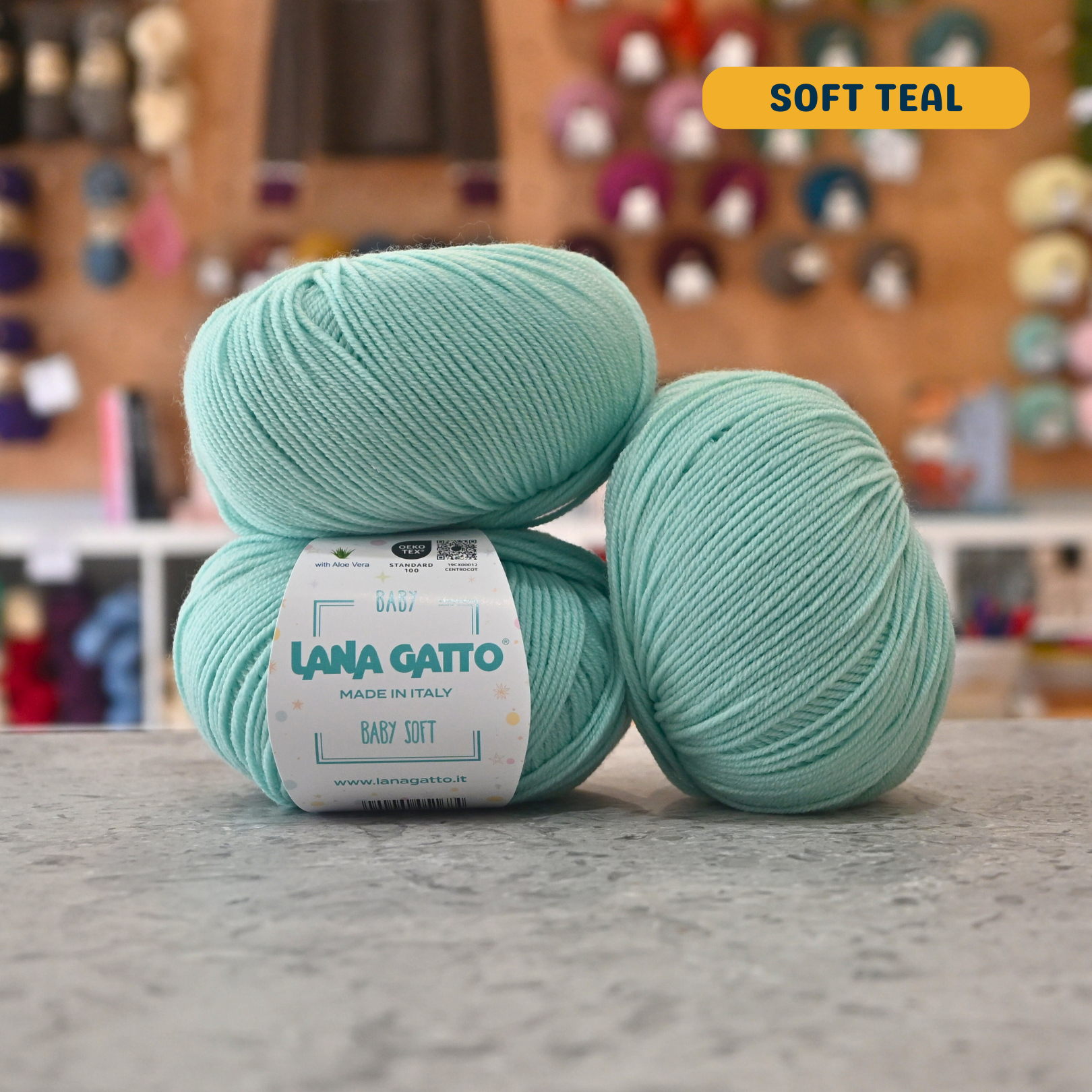 50g soft teal extrafine merino aloe vera sport yarn, 'Lana Gatto Baby Soft' label visible, craft shop background (blurred)