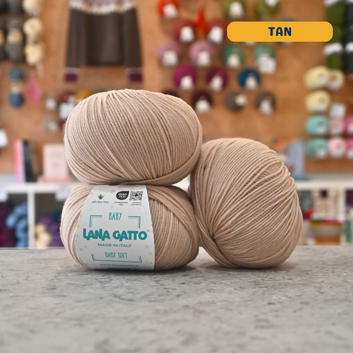 50g tan extrafine merino aloe vera sport yarn, 'Lana Gatto Baby Soft' label visible, craft shop background (blurred)
