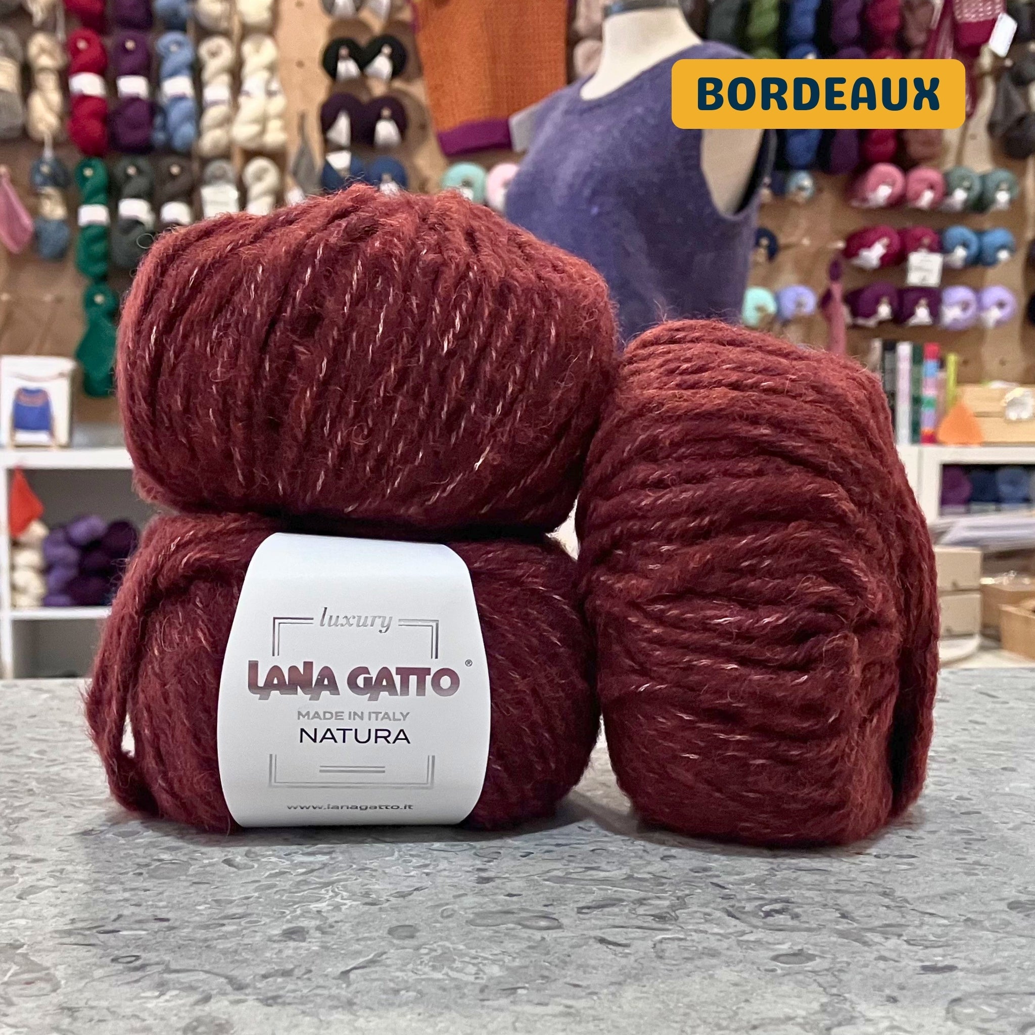 50g deep red (bordeaux) extrafine merino cotton baby alpaca bulky yarn, 'Luxury Lana Gatto Made In Italy Natura' label visible, craft shop background (blurred)
