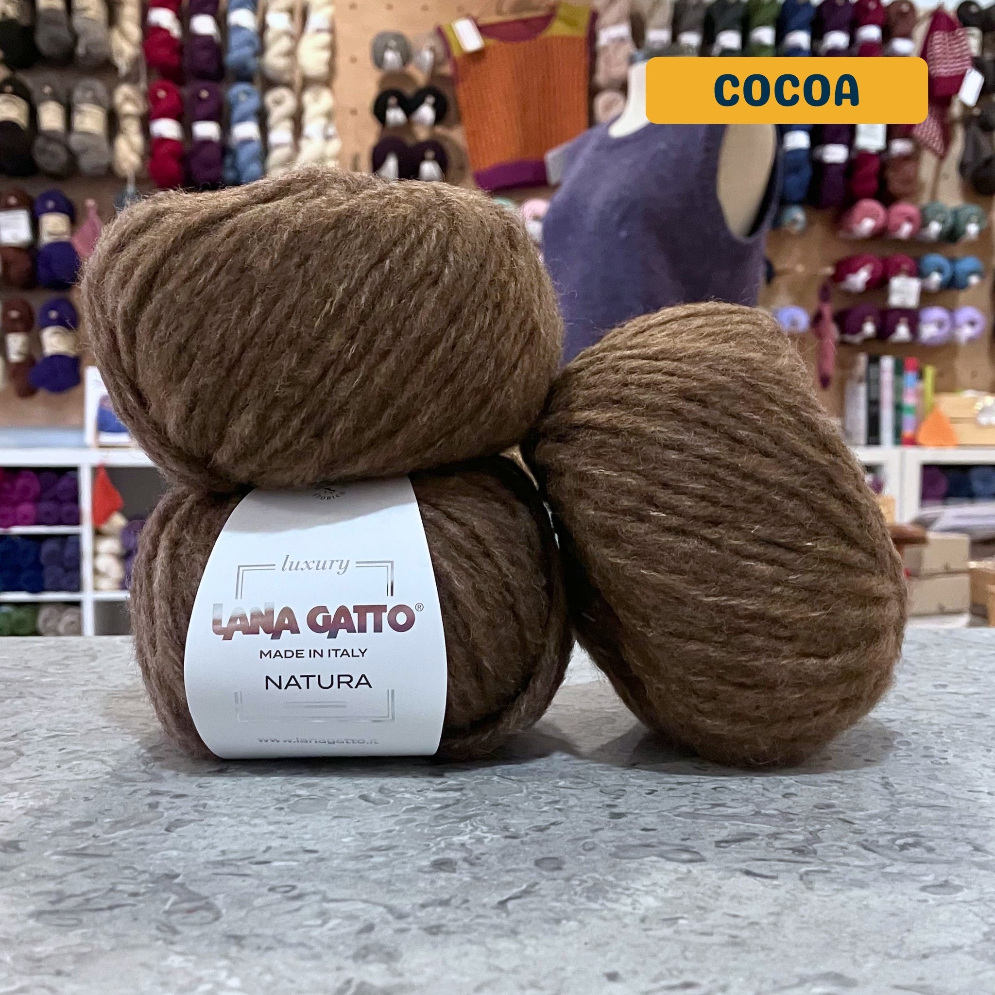 50g medium brown (cocoa) extrafine merino cotton baby alpaca bulky yarn, 'Luxury Lana Gatto Made In Italy Natura' label visible, craft shop background (blurred)