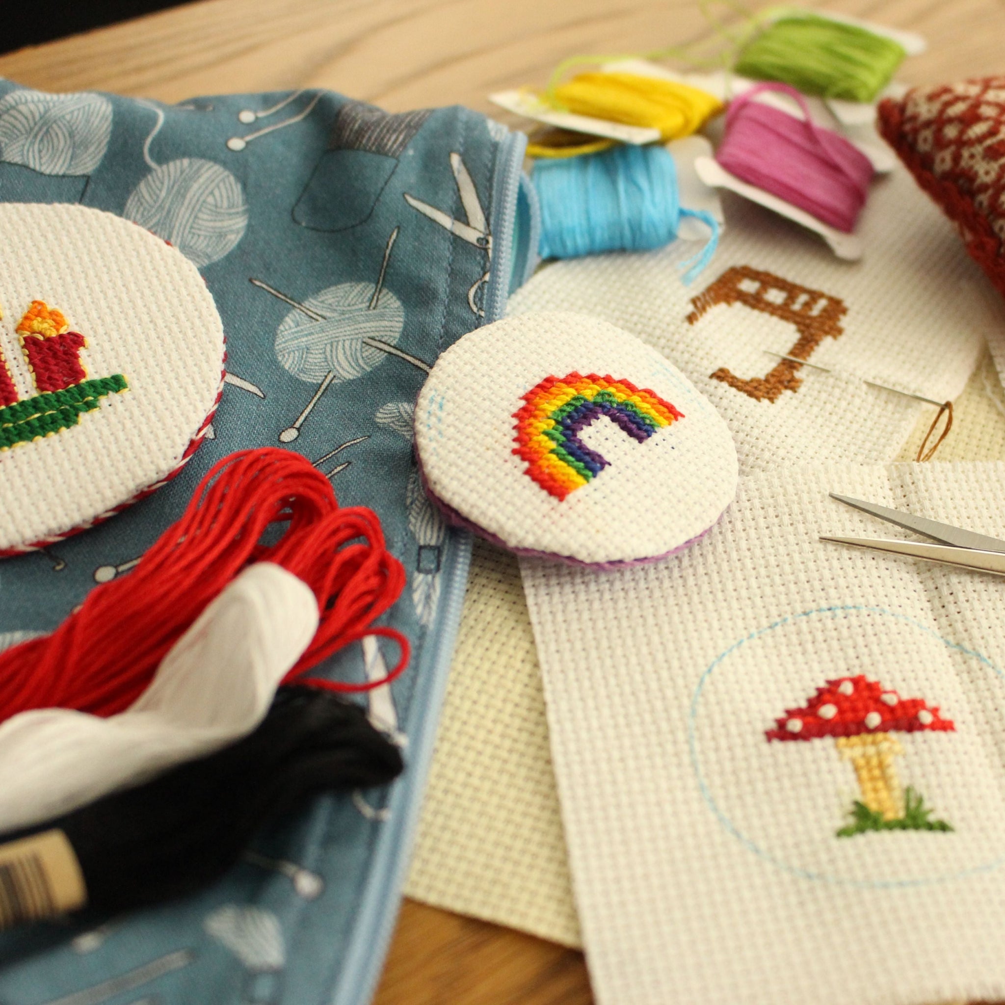 A close-up of a cross-stitch kit laid out on a wooden surface, including a blue zippered case, white Aida cloth, colorful embroidery floss, scissors, a red pincushion, and finished round cross-stitch designs featuring a rainbow and a mushroom.