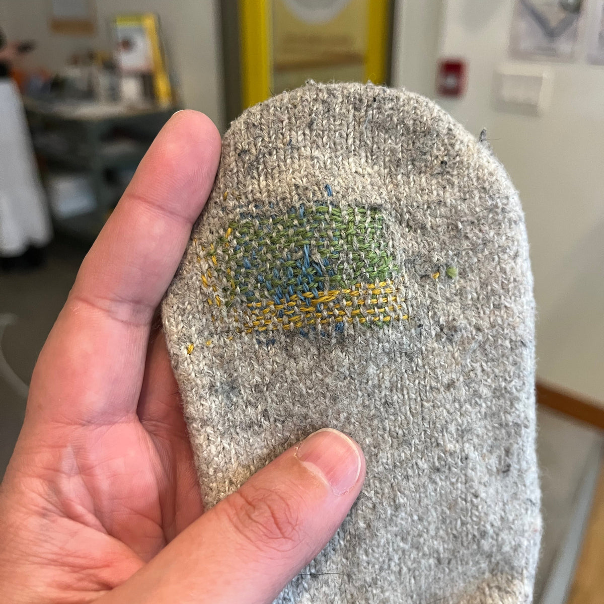 A close-up of a hand holding a gray knit sock with a colorful embroidered patch on the sole, set against a blurred indoor background.