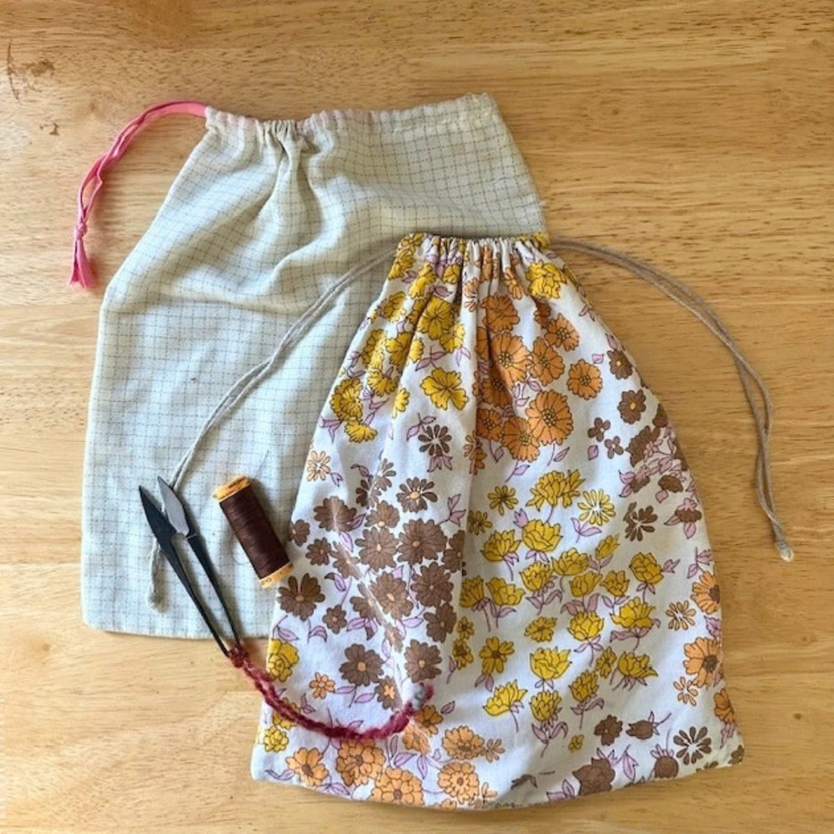 Two fabric drawstring bags laid flat on a wooden surface, with a spool of thread and thread snips. Beginner sewing workshop with Jasmine at Stitch & Knit Dartmouth — learn to sew a drawstring bag.