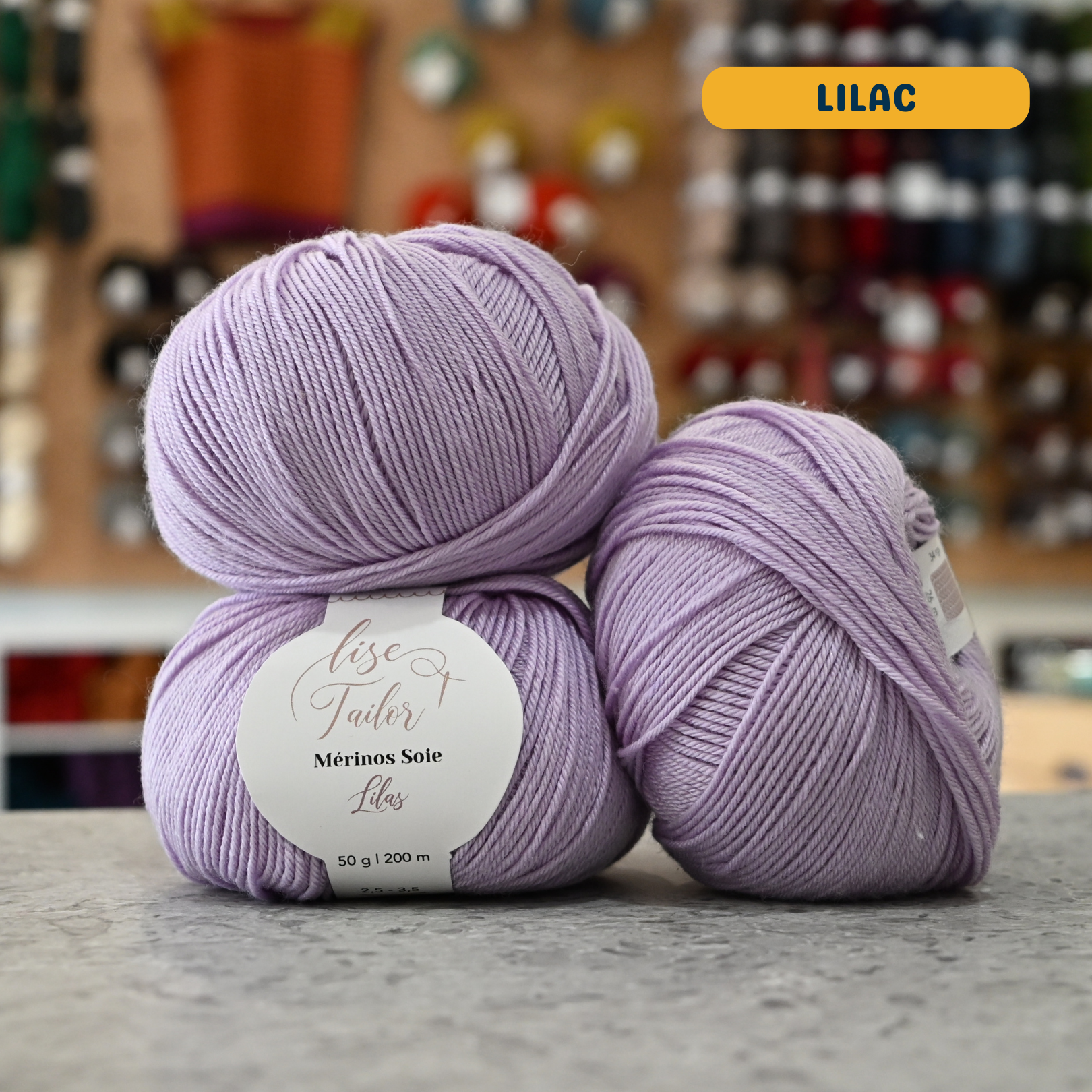 50g lilac merino & silk fingering yarn, 'Lise Tailor Merino Silk' label visible, craft shop background (blurred)