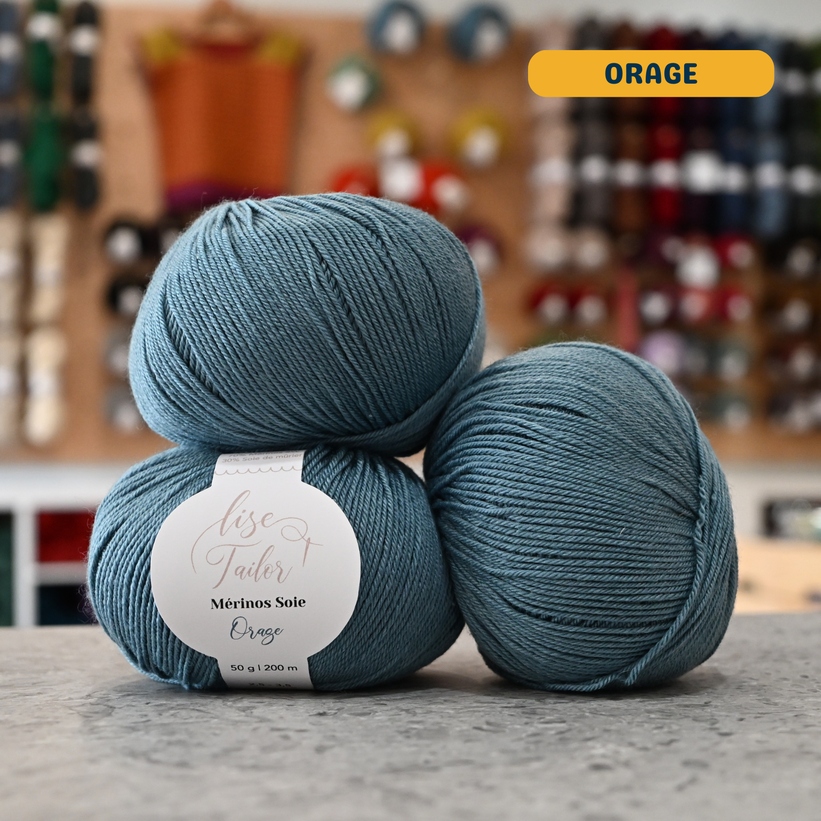 50g grey-blue (orage) merino & silk fingering yarn, 'Lise Tailor Merino Silk' label visible, craft shop background (blurred)