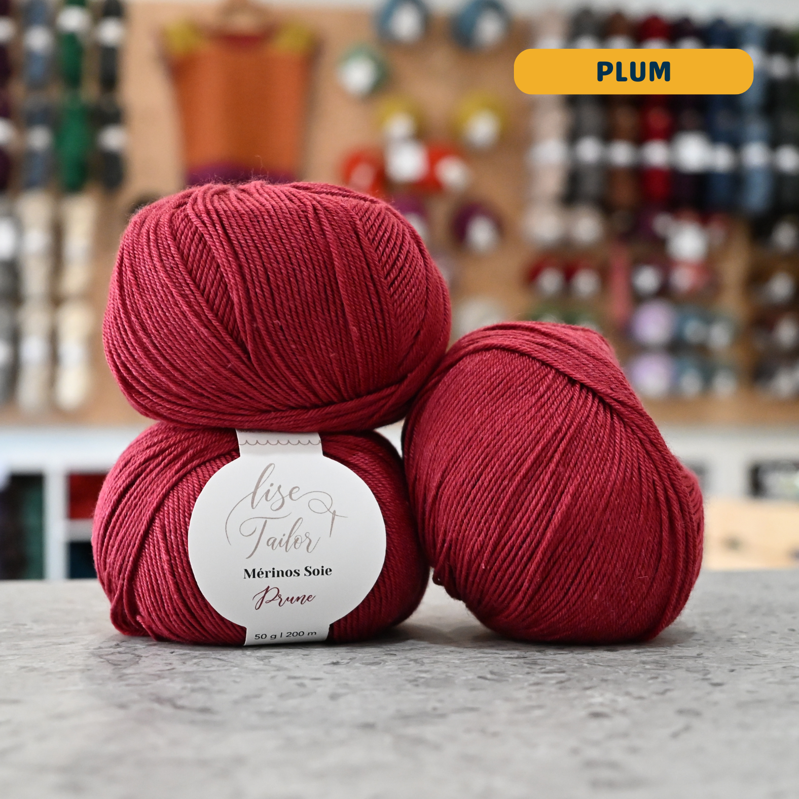 50g red (plum) merino & silk fingering yarn, 'Lise Tailor Merino Silk' label visible, craft shop background (blurred)