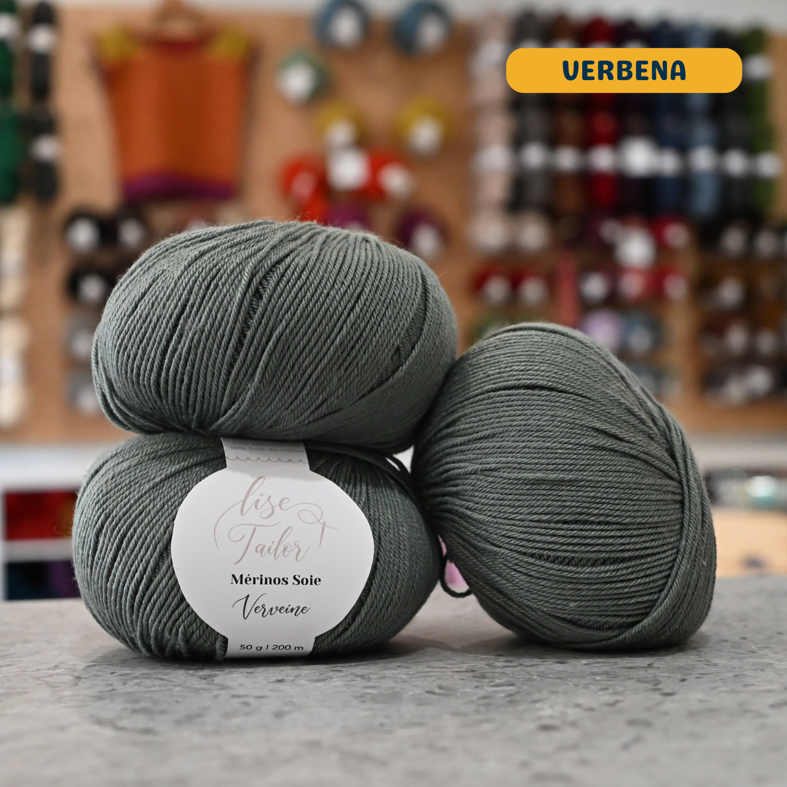 50g green-grey (verbena) merino & silk fingering yarn, 'Lise Tailor Merino Silk' label visible, craft shop background (blurred)