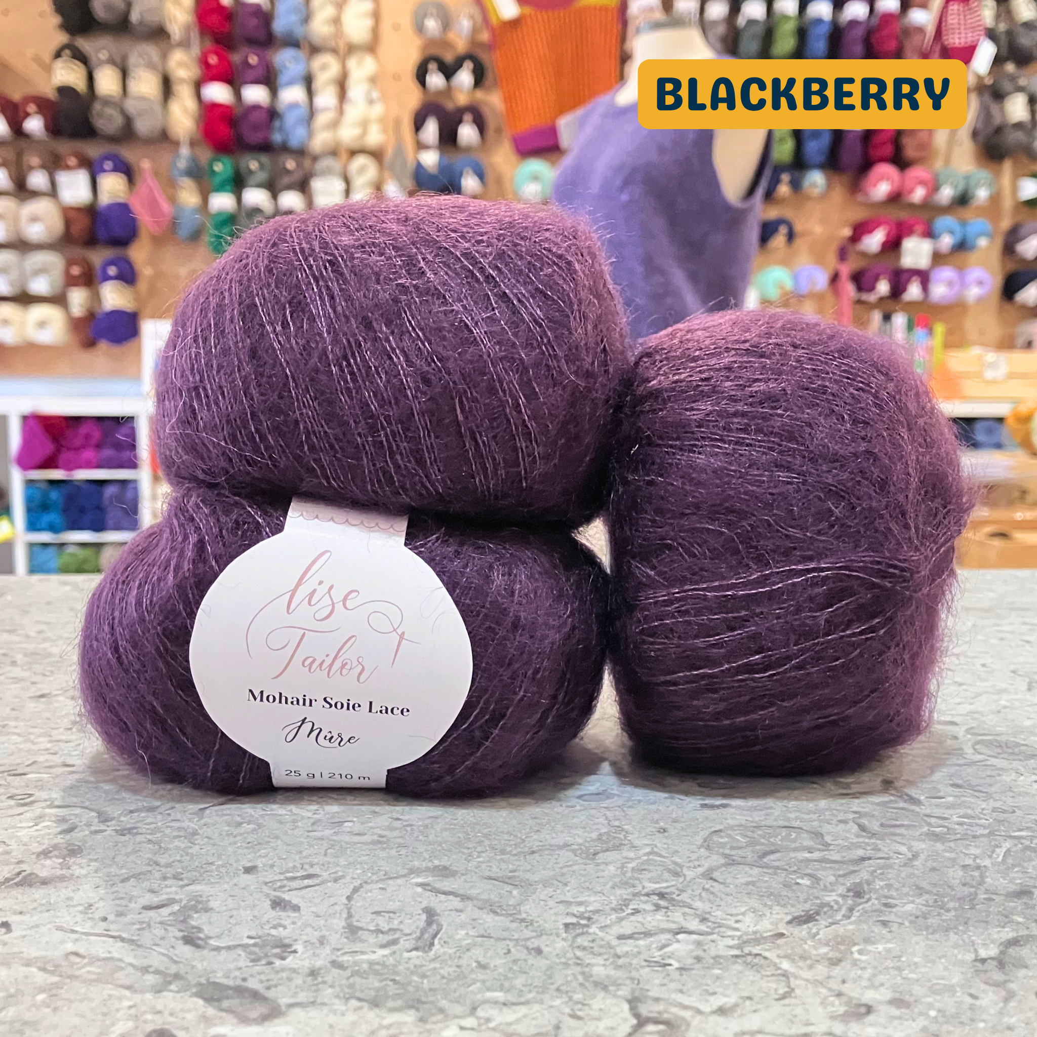 50g dark purple (blackberry) mohair & silk lace yarn, 'Lise Tailor Mohair Silk Lace' label visible, craft shop background (blurred)
