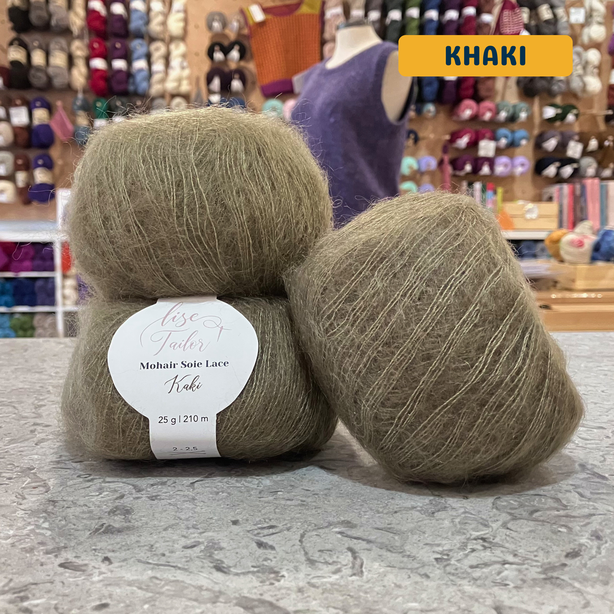 50g green-grey-brown (khaki) mohair & silk lace yarn, 'Lise Tailor Mohair Silk Lace' label visible, craft shop background (blurred)
