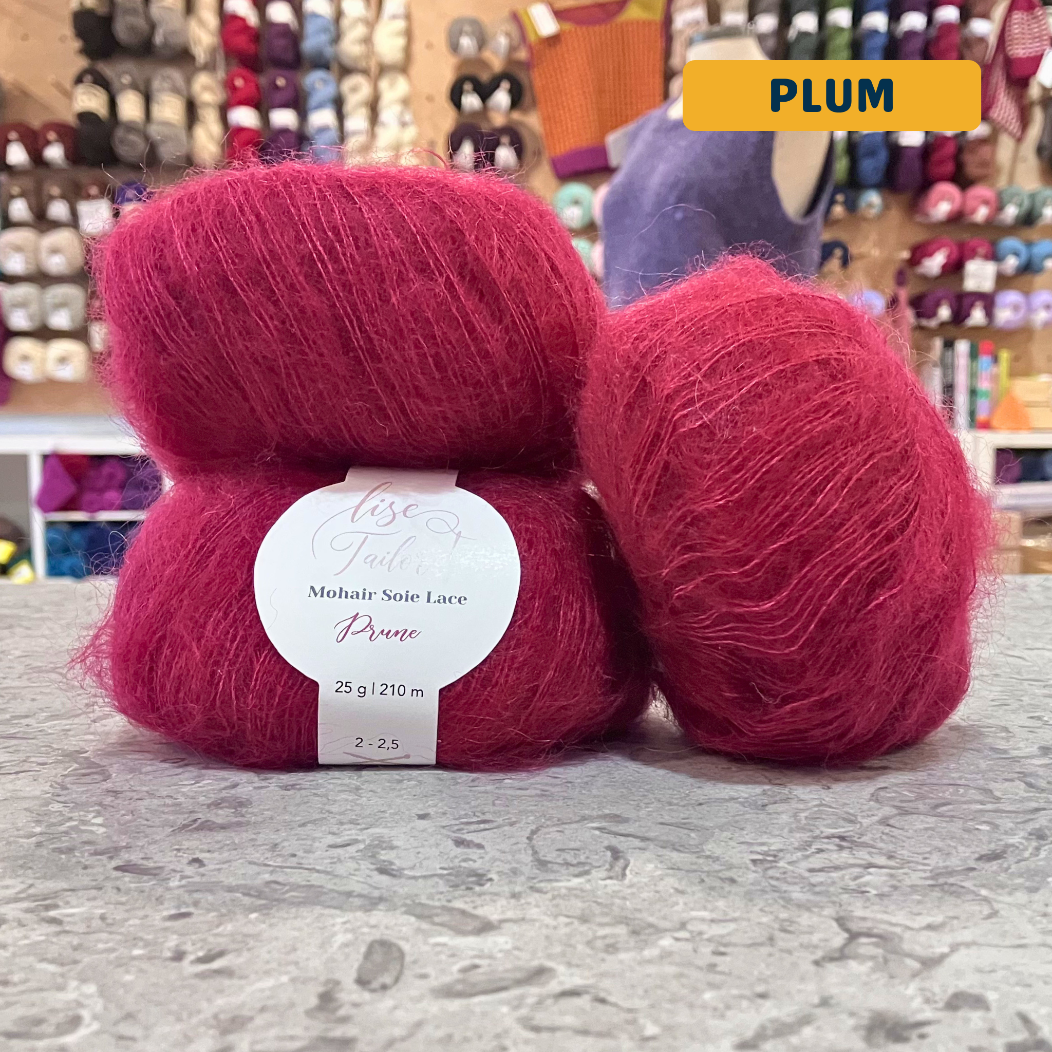 50g red (plum) mohair & silk lace yarn, 'Lise Tailor Mohair Silk Lace' label visible, craft shop background (blurred)