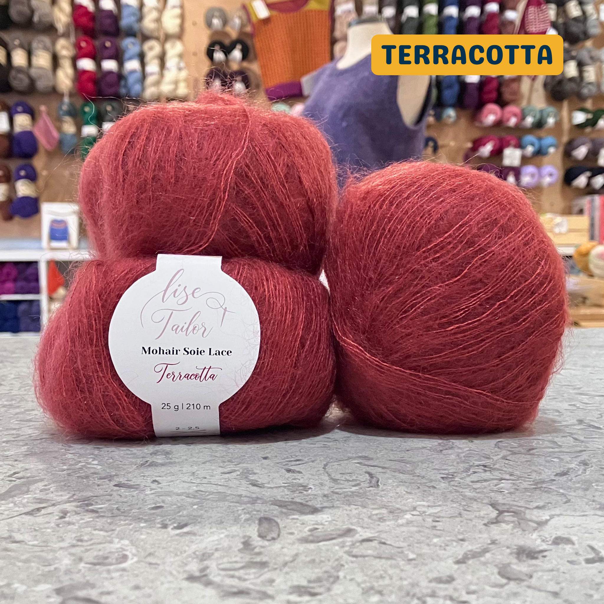 50g orange-red (terracotta) mohair & silk lace yarn, 'Lise Tailor Mohair Silk Lace' label visible, craft shop background (blurred)
