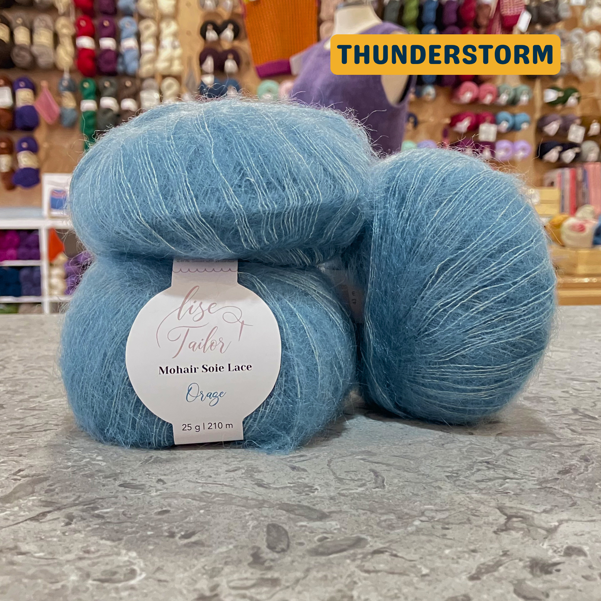 50g grey-turquoise (thunderstorm) mohair & silk lace yarn, 'Lise Tailor Mohair Silk Lace' label visible, craft shop background (blurred)